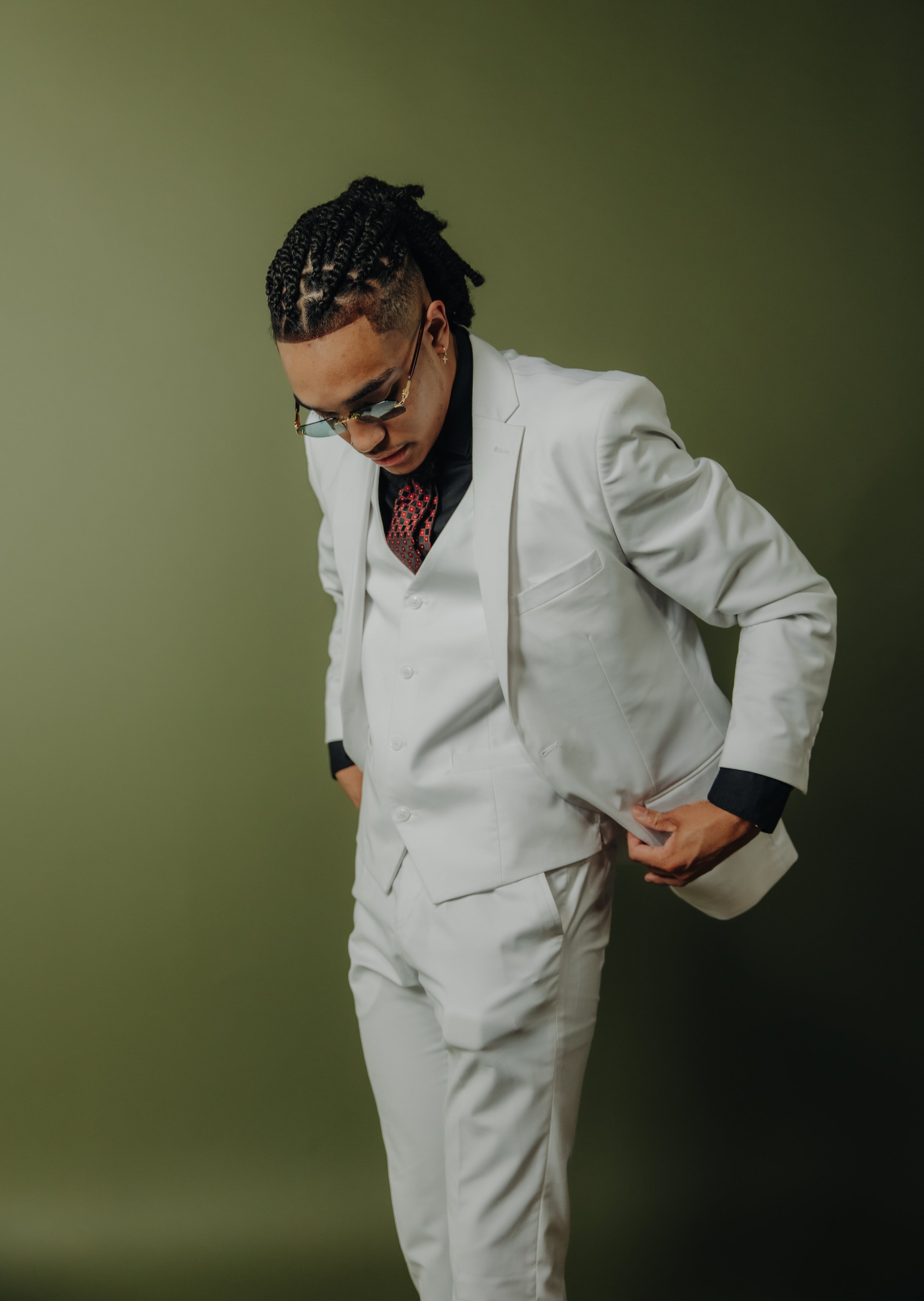 Person in a white suit with braids and sunglasses, adjusting the jacket, set against a green background.