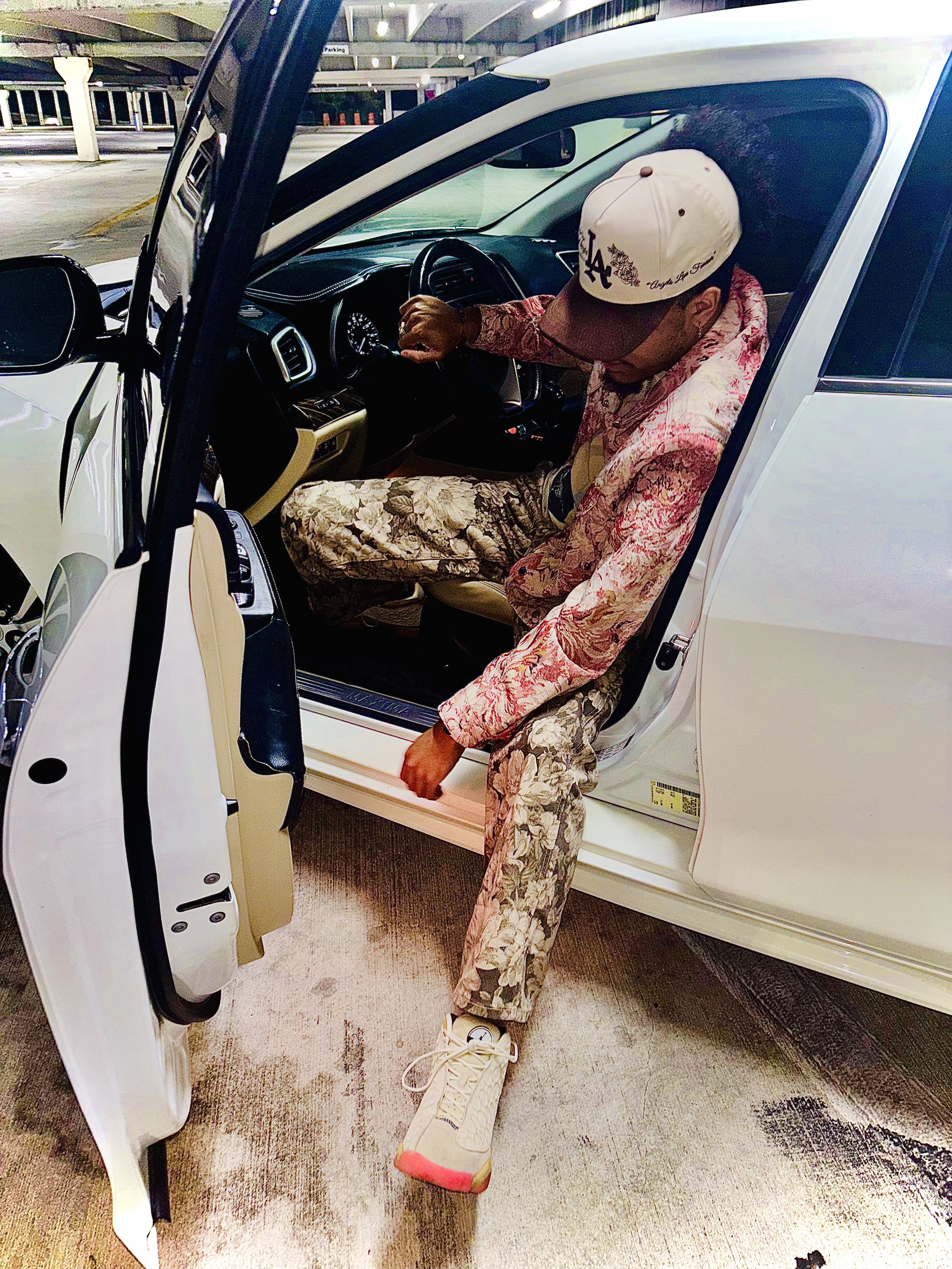 Person wearing patterned clothing and a baseball cap sitting in a car with an open door in a parking garage.