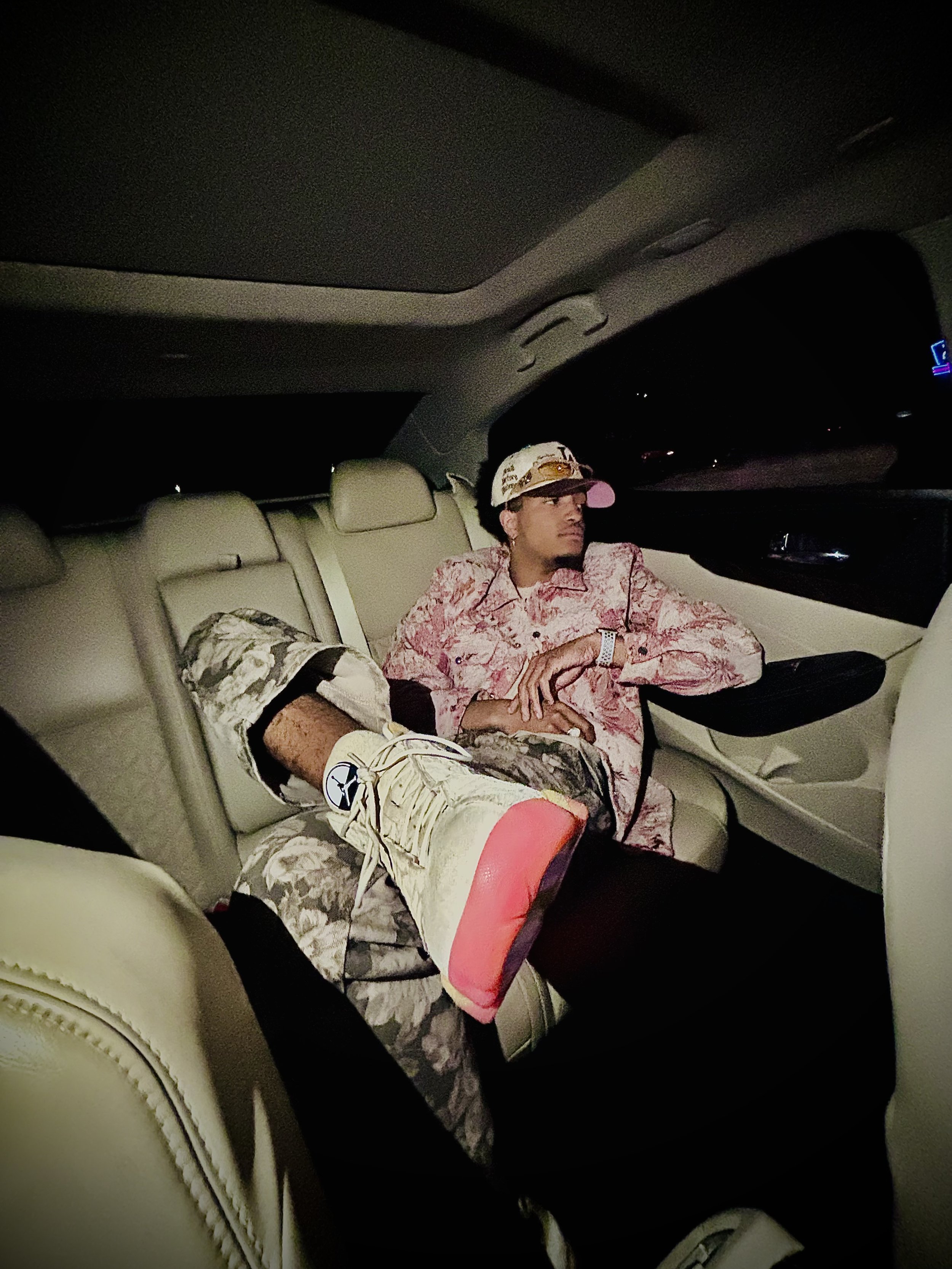 Man in camouflage pants and pink shirt reclining in a car interior, wearing a cap and sneakers.