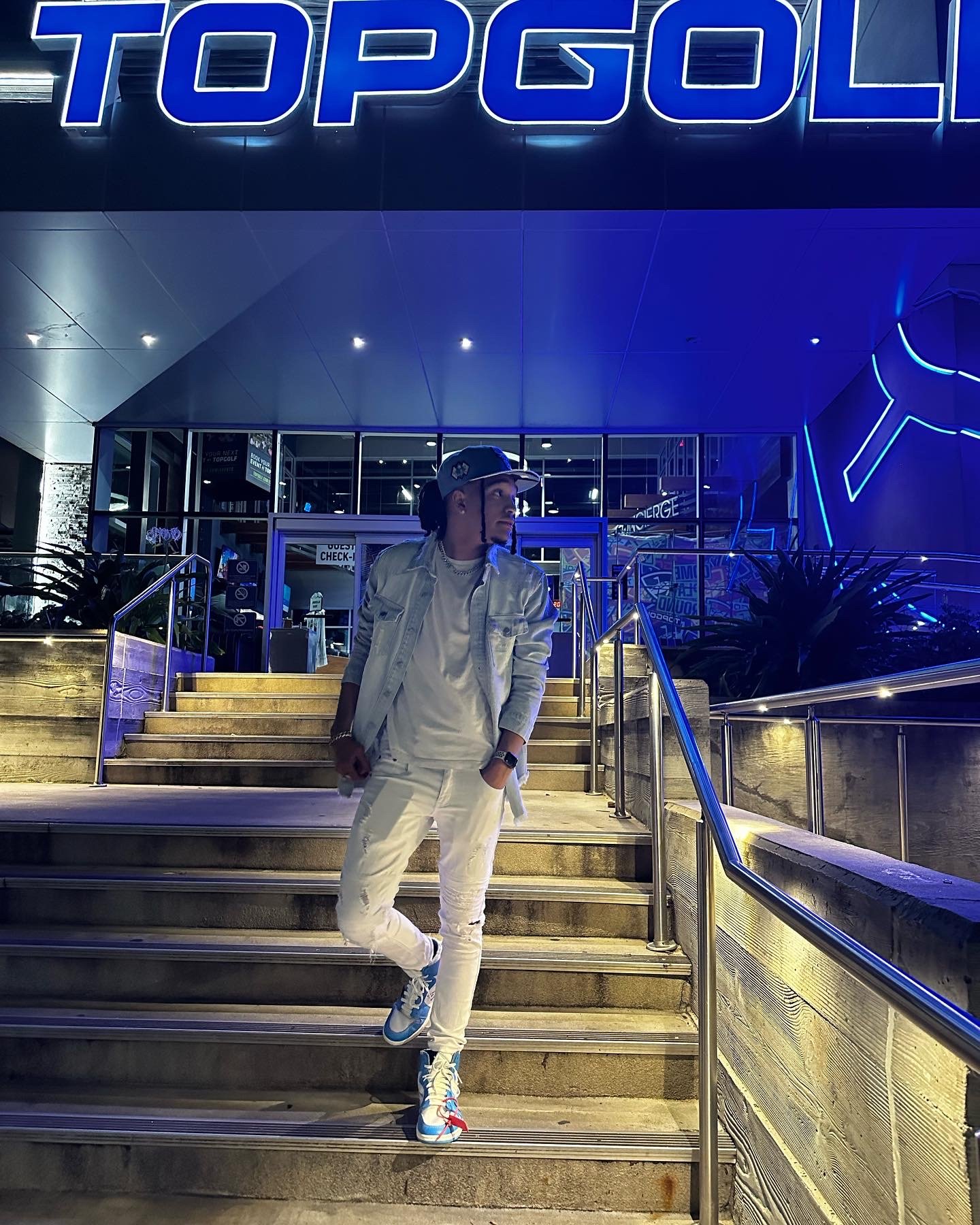 Person standing on stairs outside Topgolf at night, wearing white clothing and blue sneakers, under blue neon lighting.