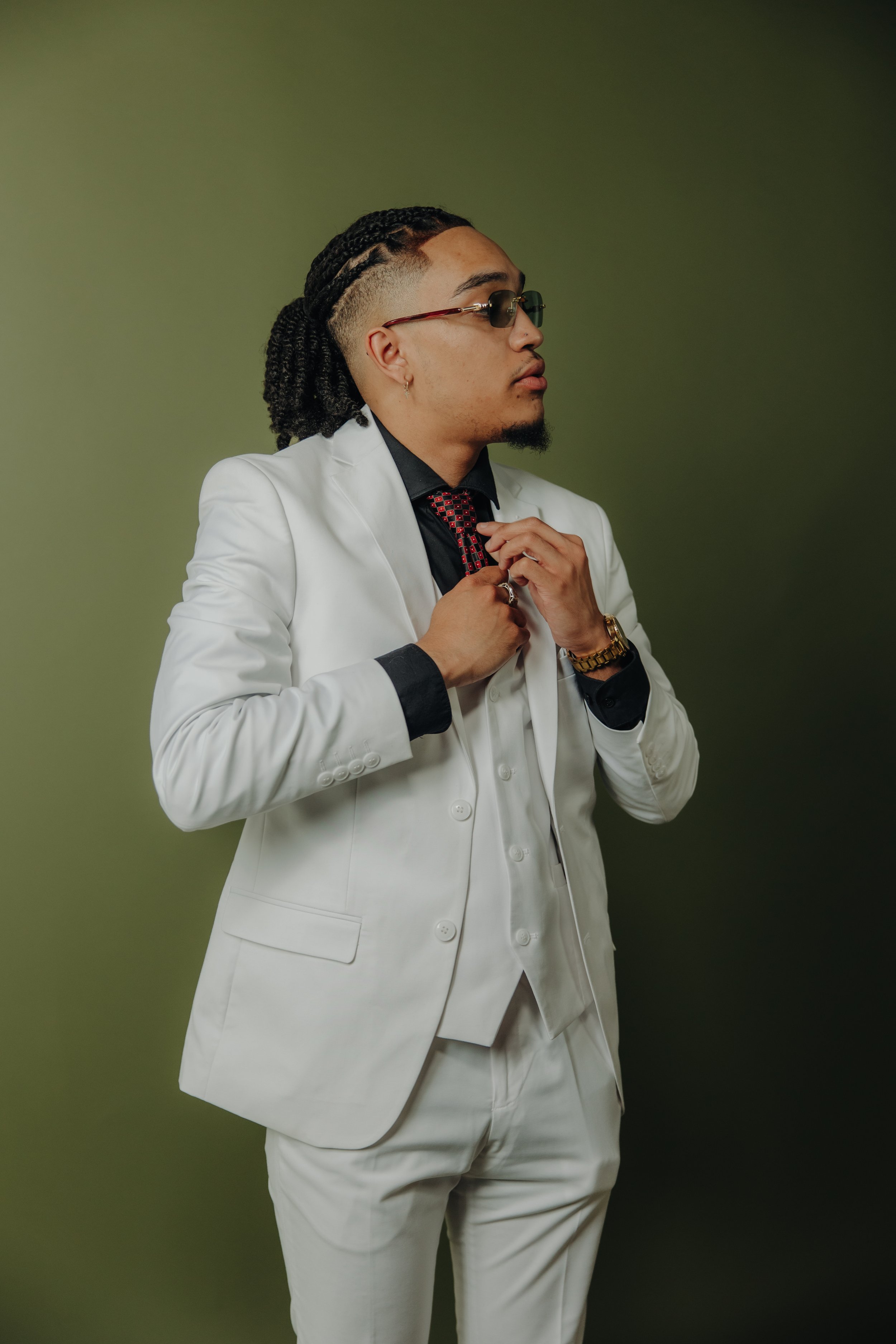Man in a white suit adjusting his red tie against a green background.