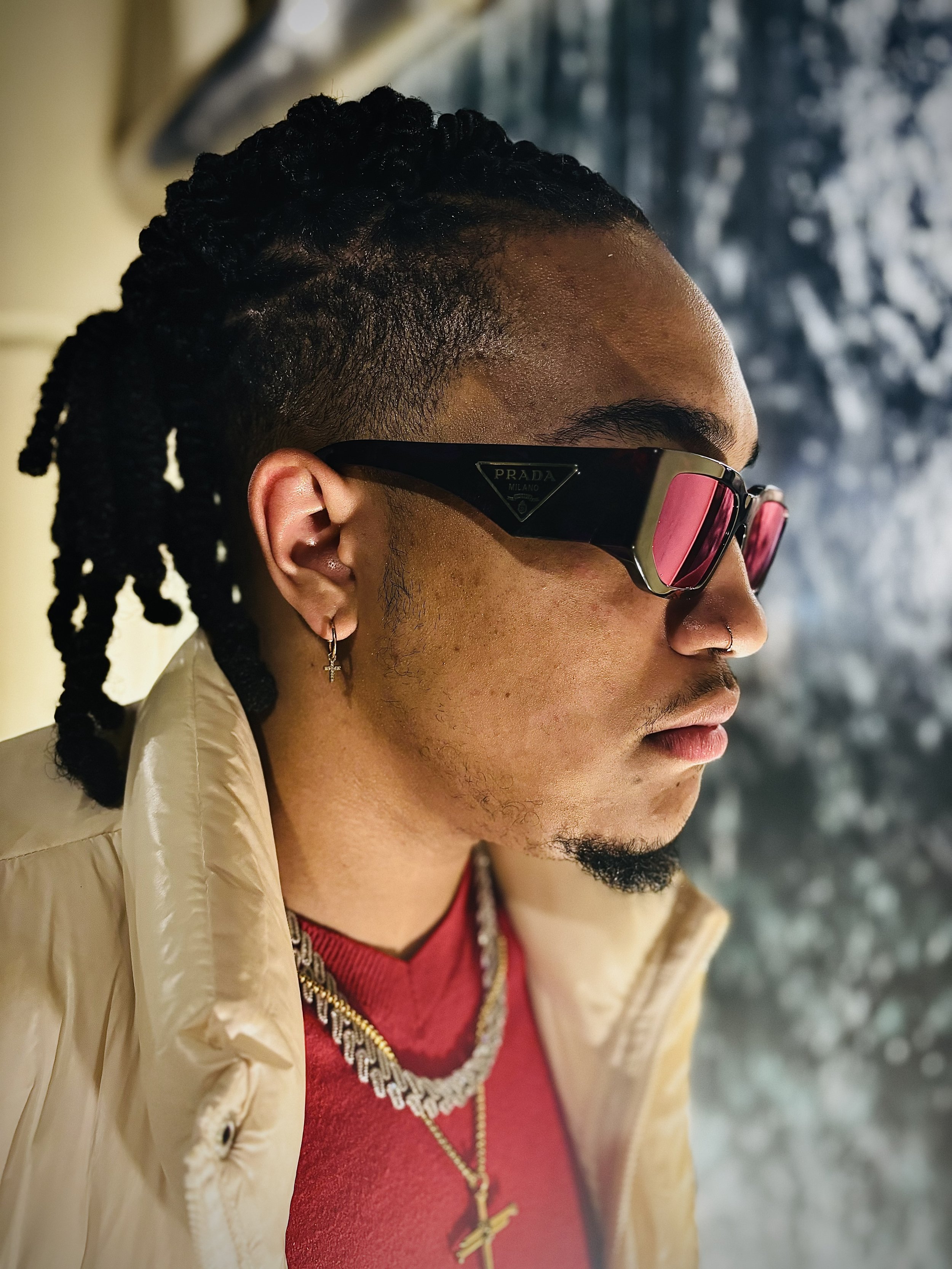 Side profile of a person wearing sunglasses, a beige puffer jacket, a red shirt, and jewelry.