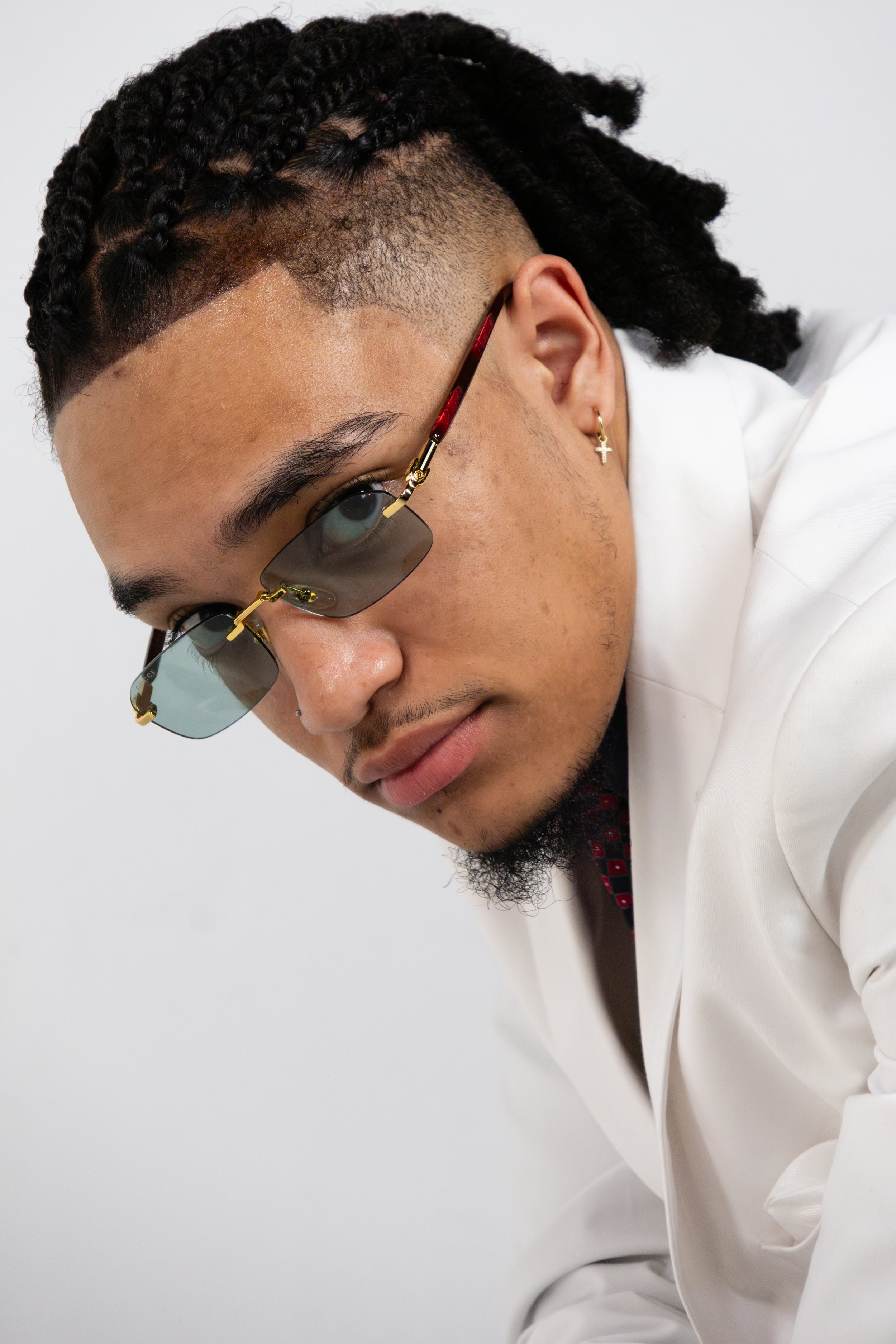 Close-up of a man in a white suit with cornrow braids, sunglasses, and a gold cross earring, looking at the camera.