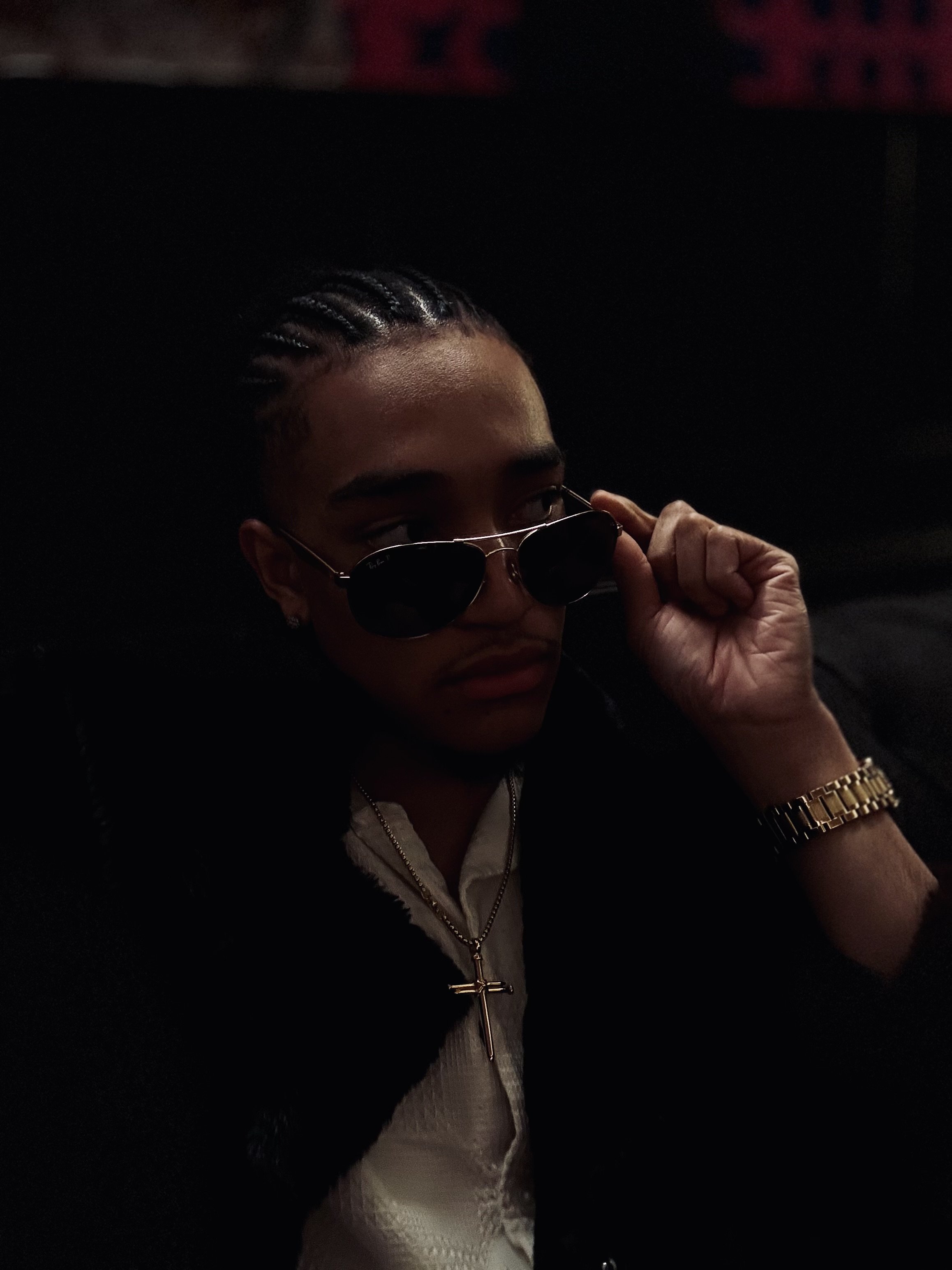 Person wearing sunglasses and jewelry, adjusting glasses, dark background.