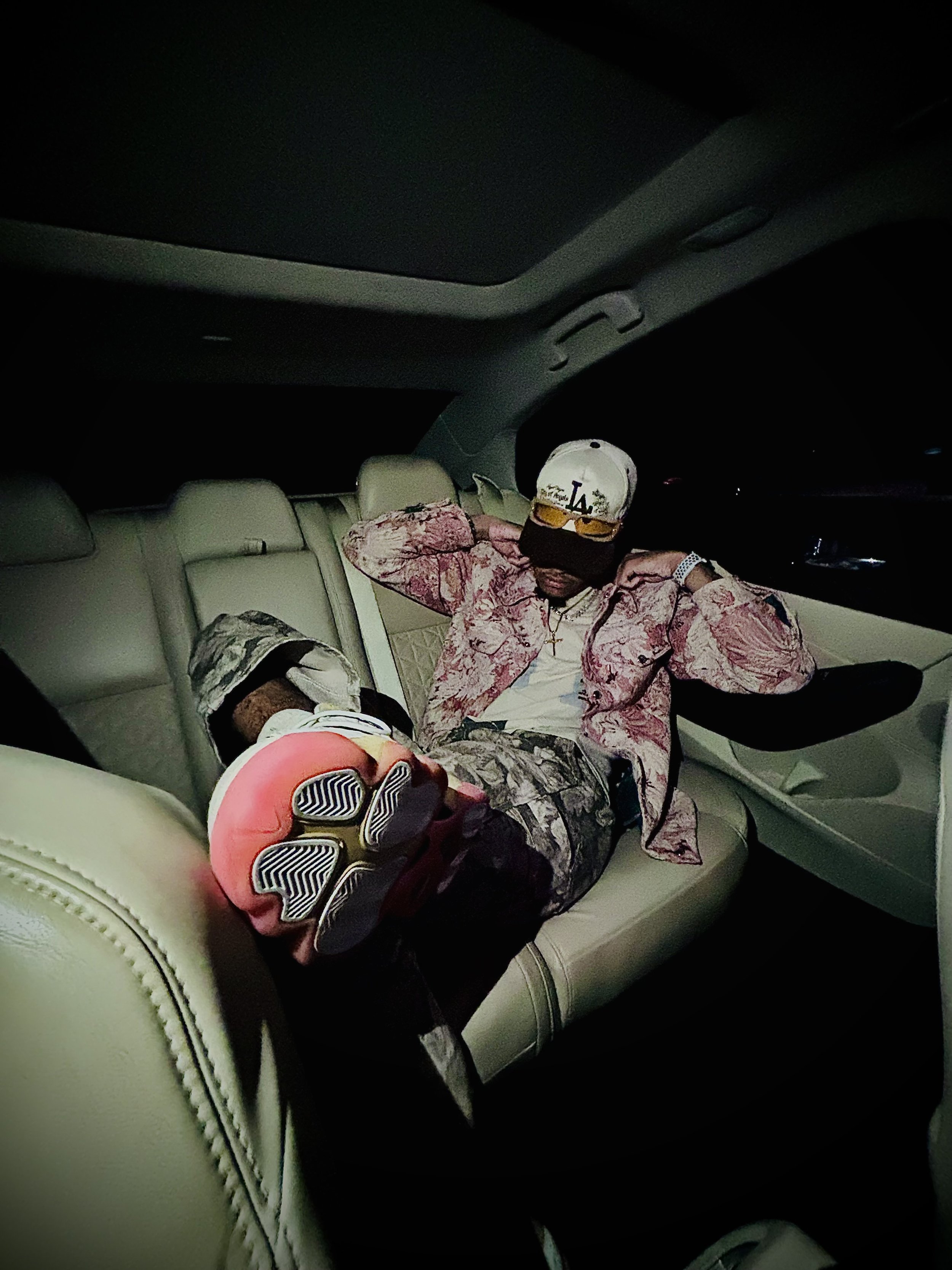 Person sitting in the backseat of a car, wearing a patterned jacket, camo pants, a cap, sunglasses, and colorful sneakers with a visible tread. The car interior is dimly lit.