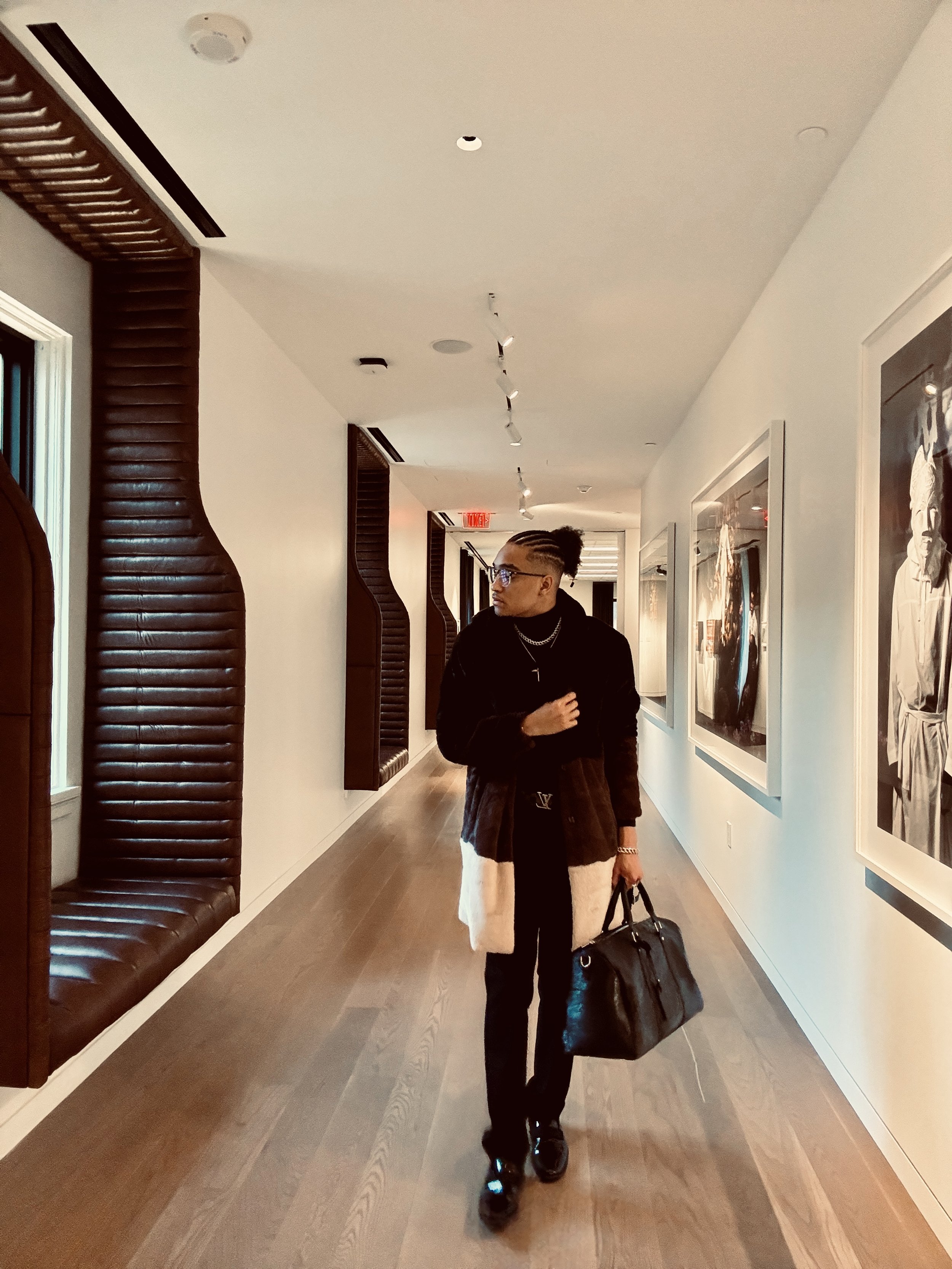 Person walking in a modern hallway with framed artwork, wearing a coat and carrying a black bag.