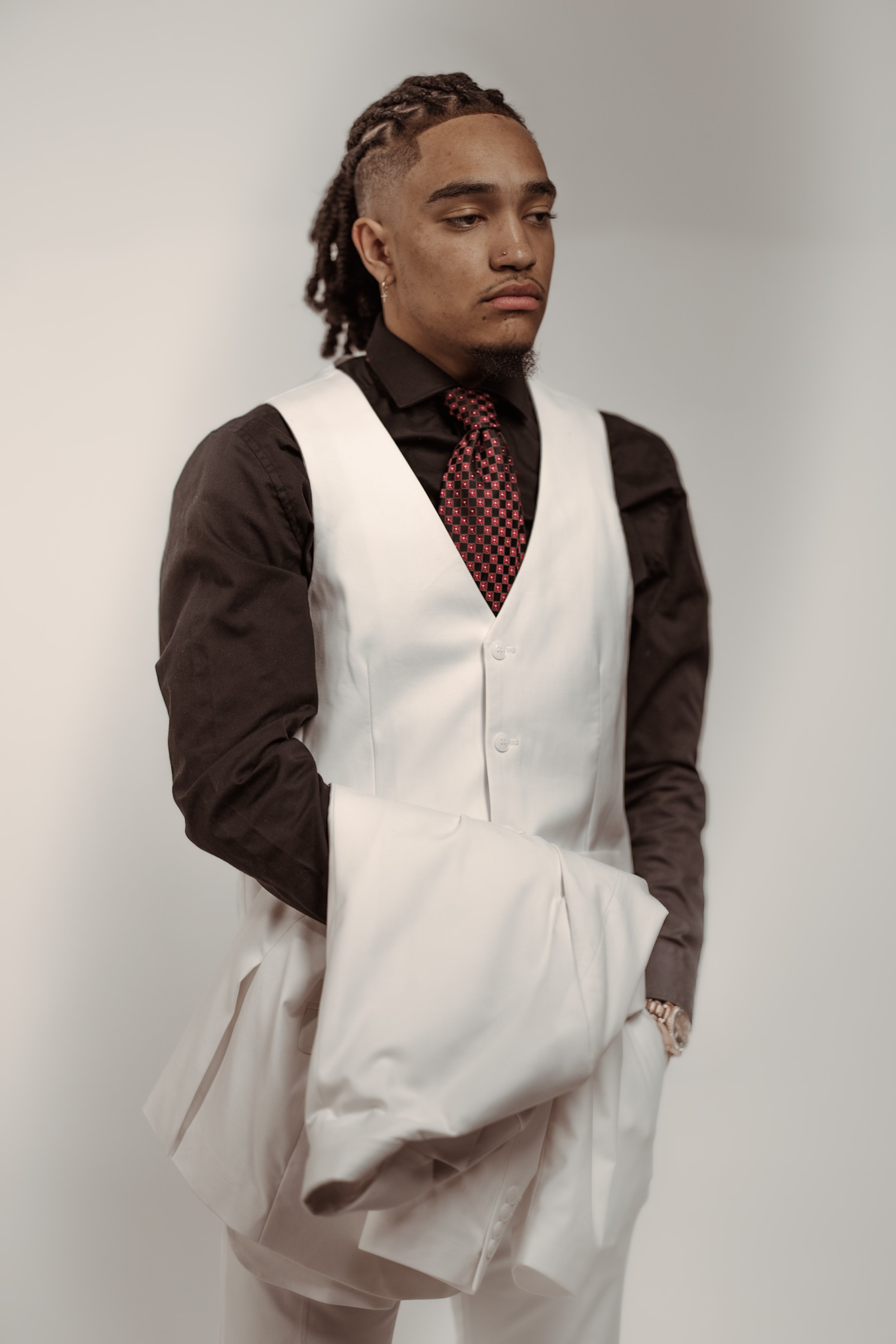 A person wearing a white vest and black shirt with a checkered tie, holding a white jacket over their arm. The individual has braided hair and is standing against a neutral background.