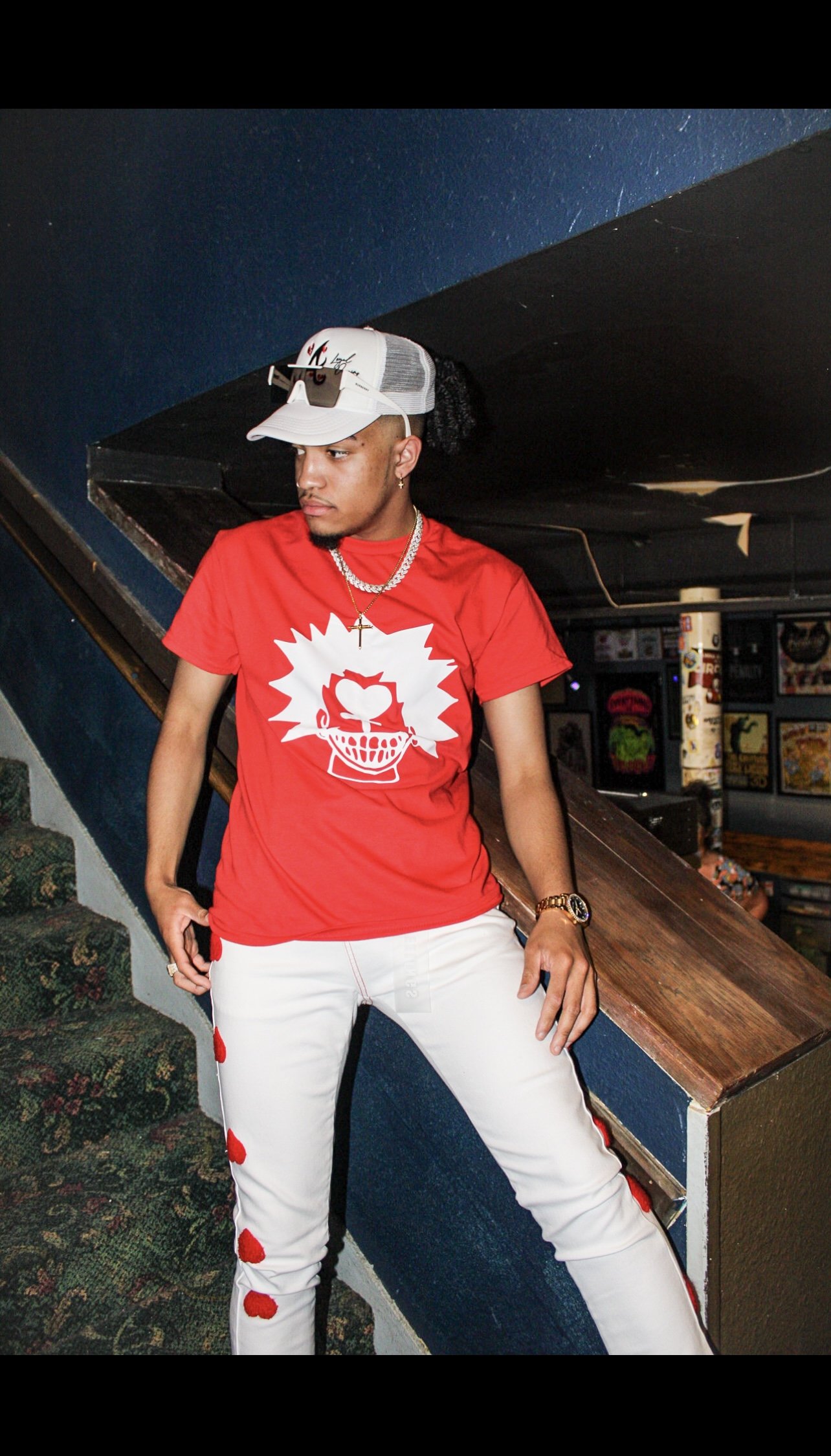A person wearing a red t-shirt with a graphic design, white pants with red patches, a cap, sunglasses on top, and a necklace, standing on a staircase in a dimly lit room.