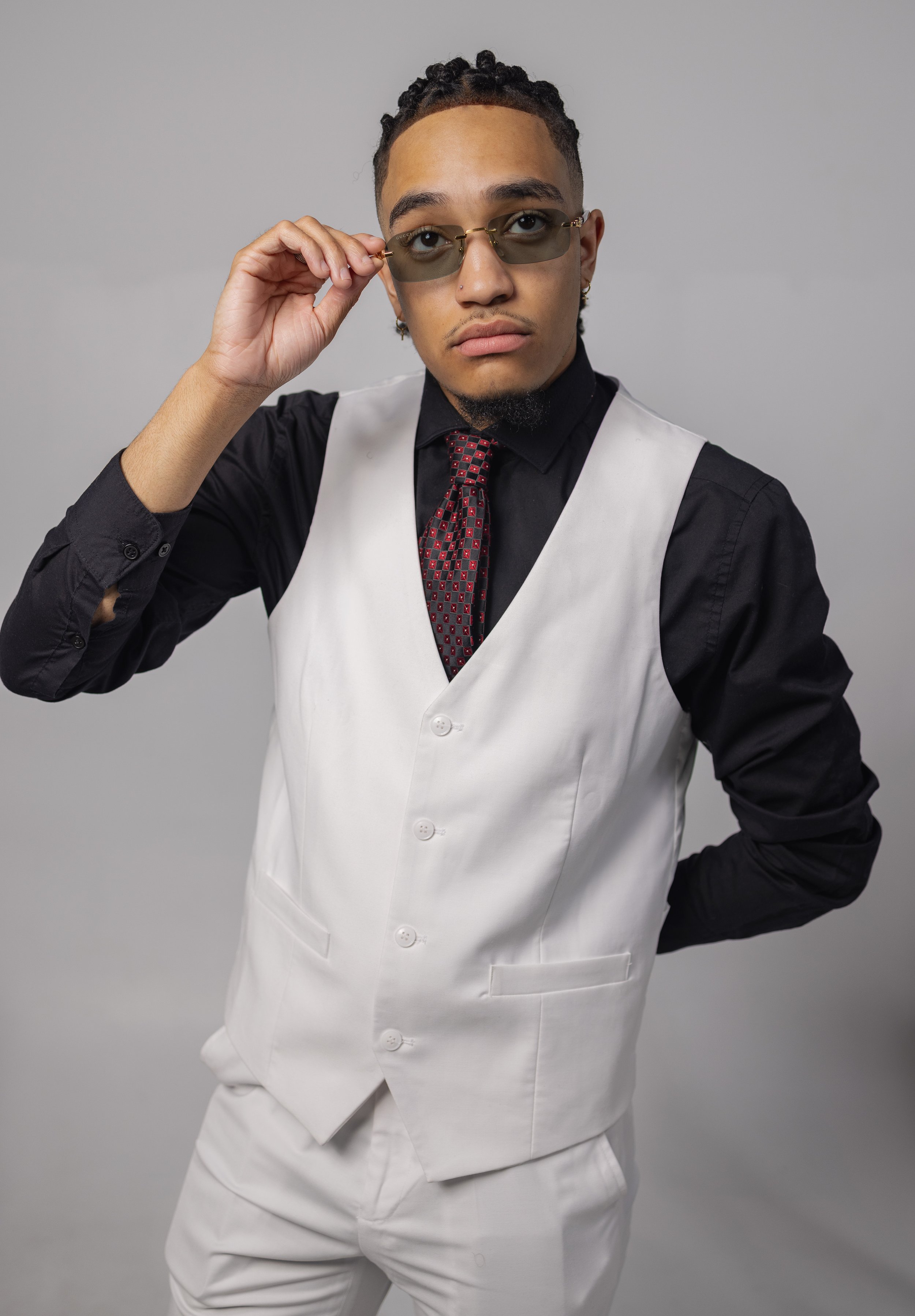 Man in a stylish white suit with a black shirt and red tie, wearing sunglasses, holding one arm behind his back and adjusting his glasses with the other hand.