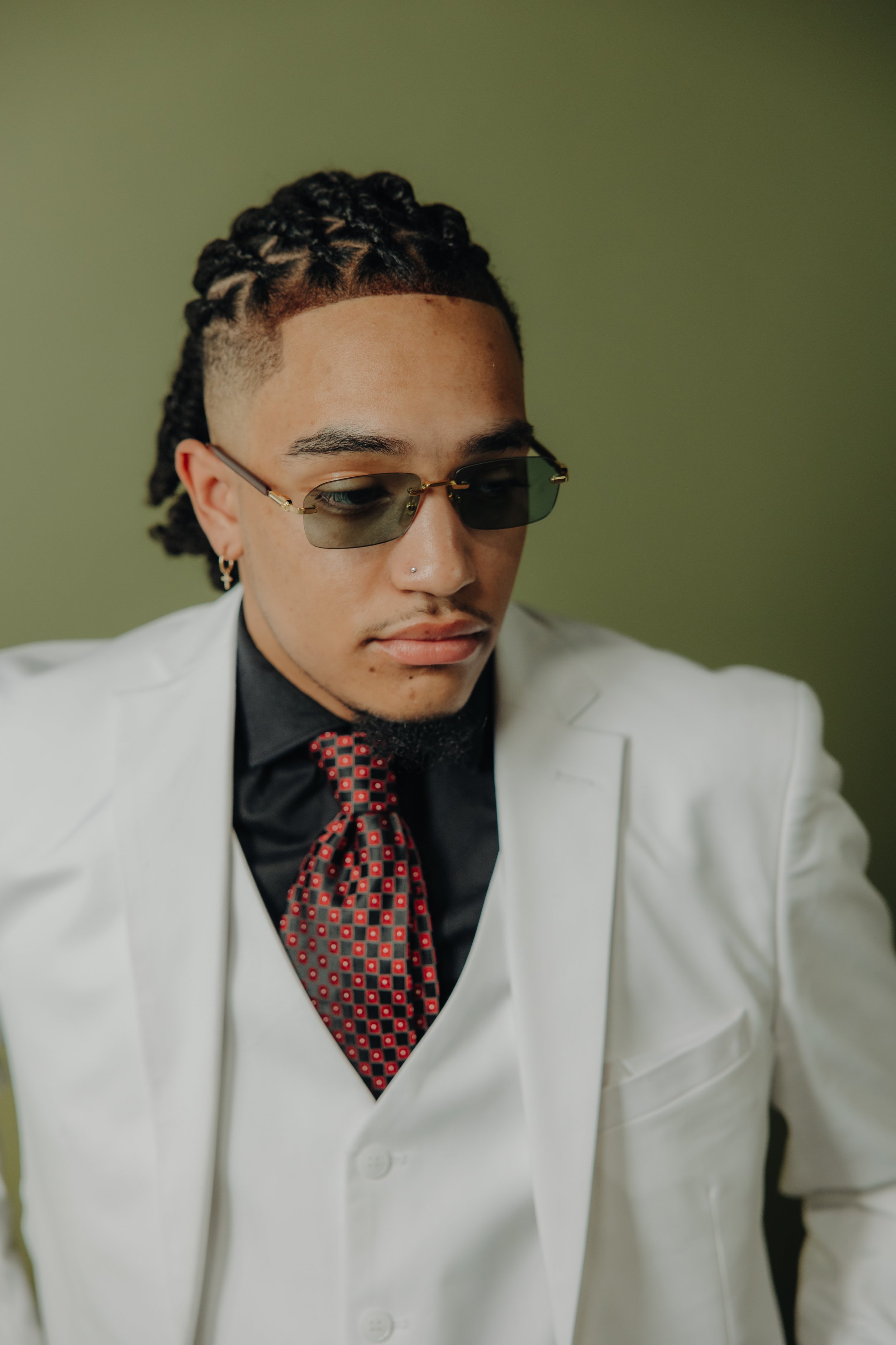 Man wearing a white suit, black shirt, patterned red tie, and sunglasses, with braided hairstyle, against a green background.