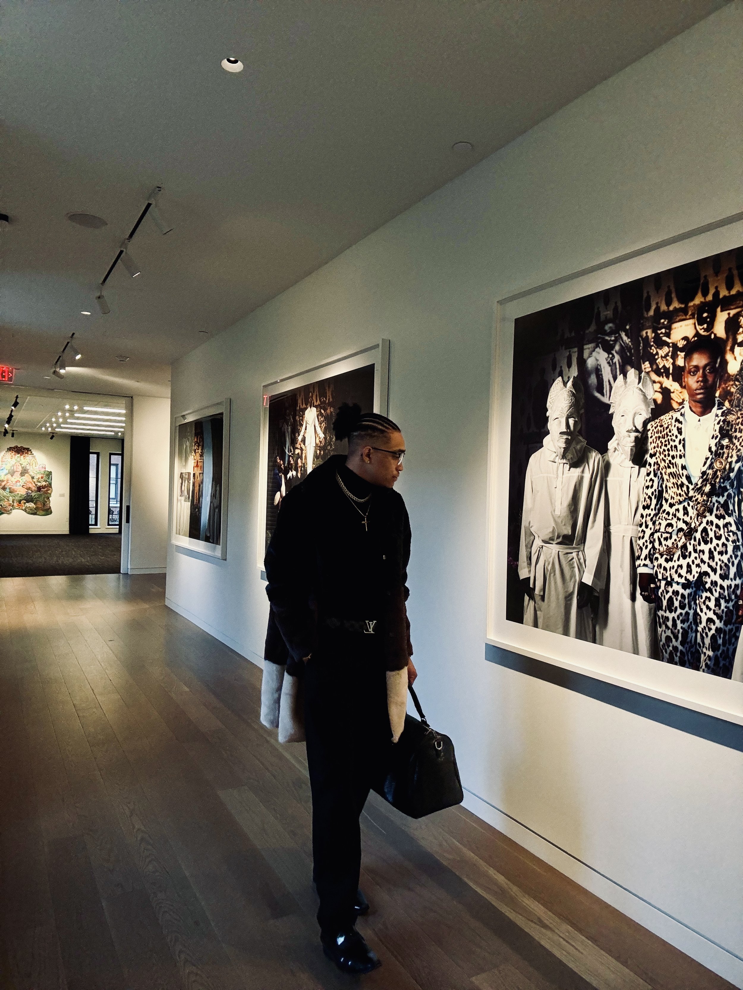 Person walking through an art gallery, observing large framed photographs on the wall. The gallery has wooden floors and track lighting, and the individual carries a black bag, wearing dark clothing.