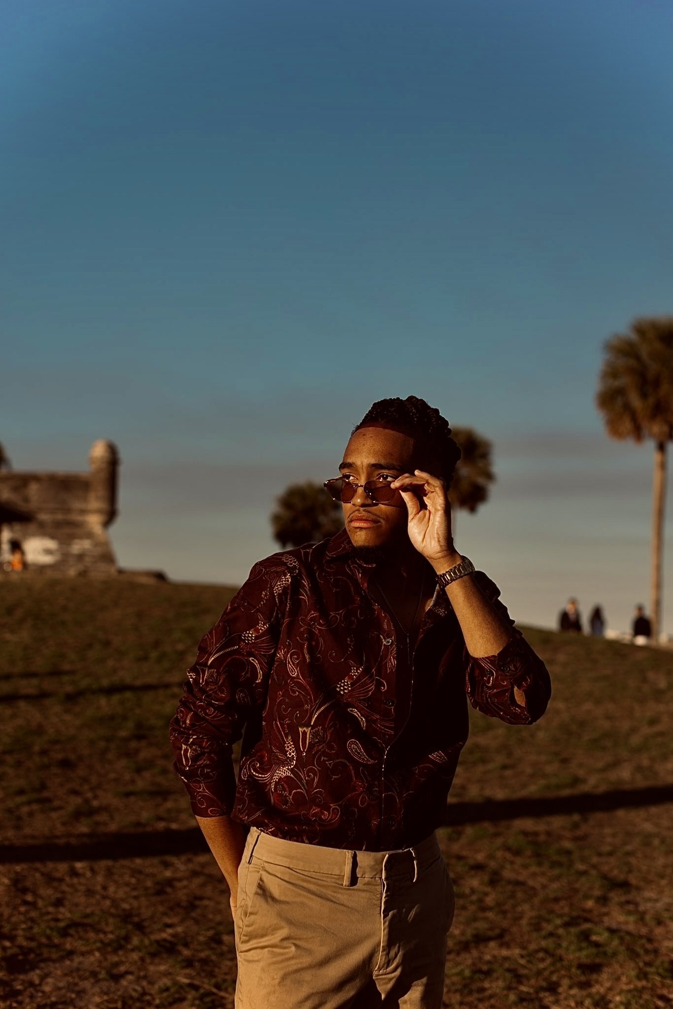 Person outdoors wearing sunglasses, patterned shirt, and khaki pants, with palm trees and an old stone building in the background.