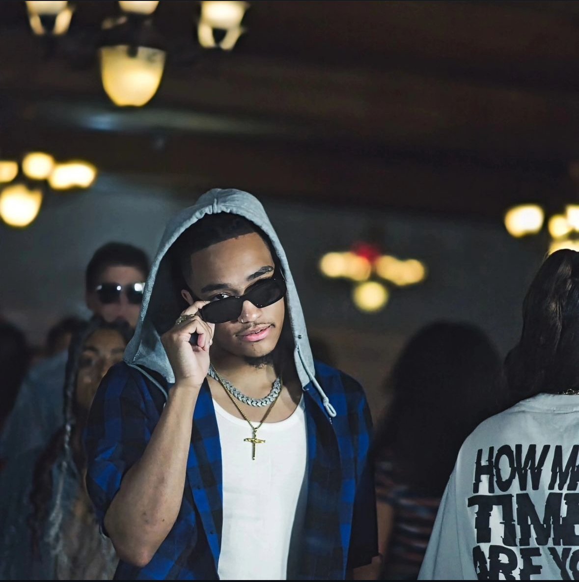A young man wearing sunglasses, a plaid shirt over a hoodie, and a necklace with a cross pendant in a dimly lit room.