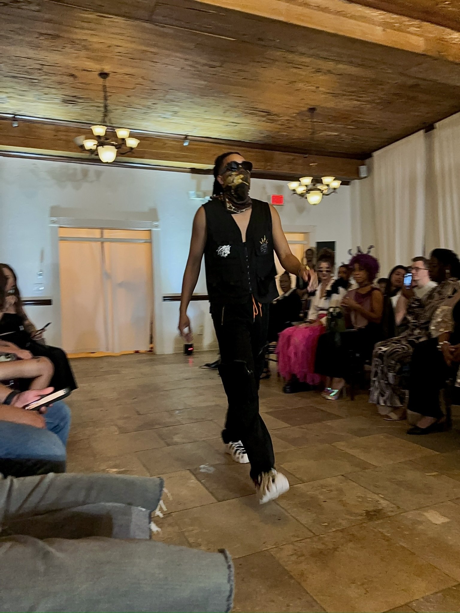 A model walking down a runway in a dimly lit room, wearing a sleeveless black outfit and a face covering, with an audience seated on both sides.