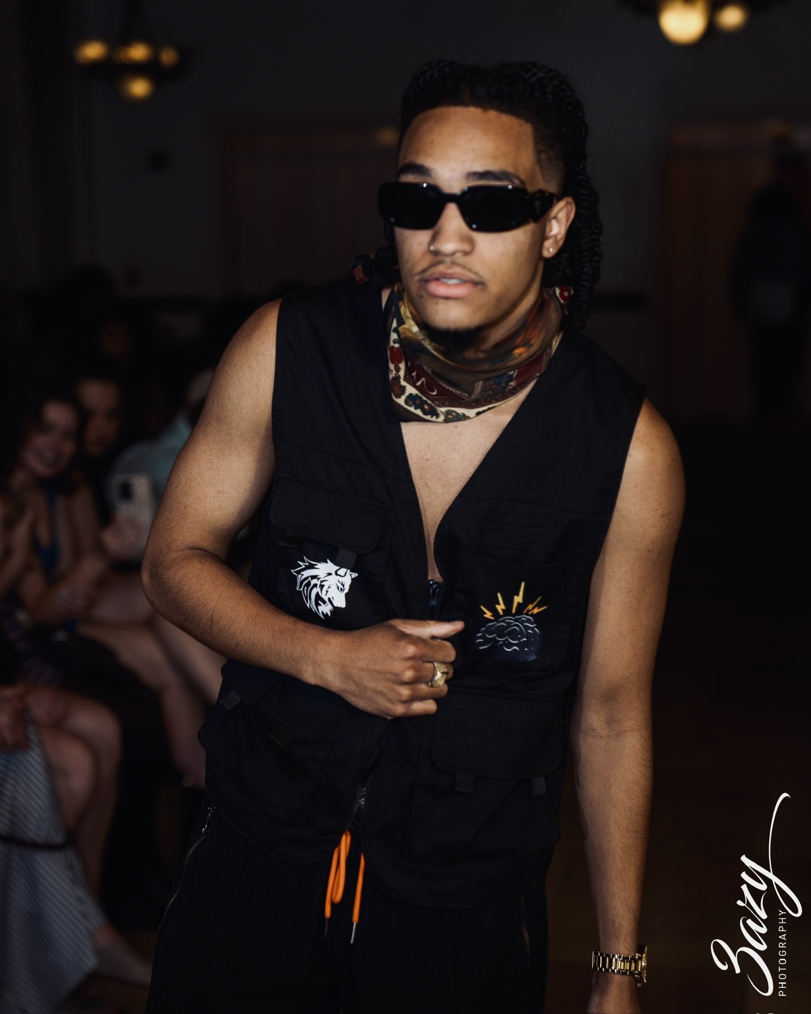 A person wearing sunglasses and a sleeveless jacket with designs, standing in a dimly lit setting, with people in the background.