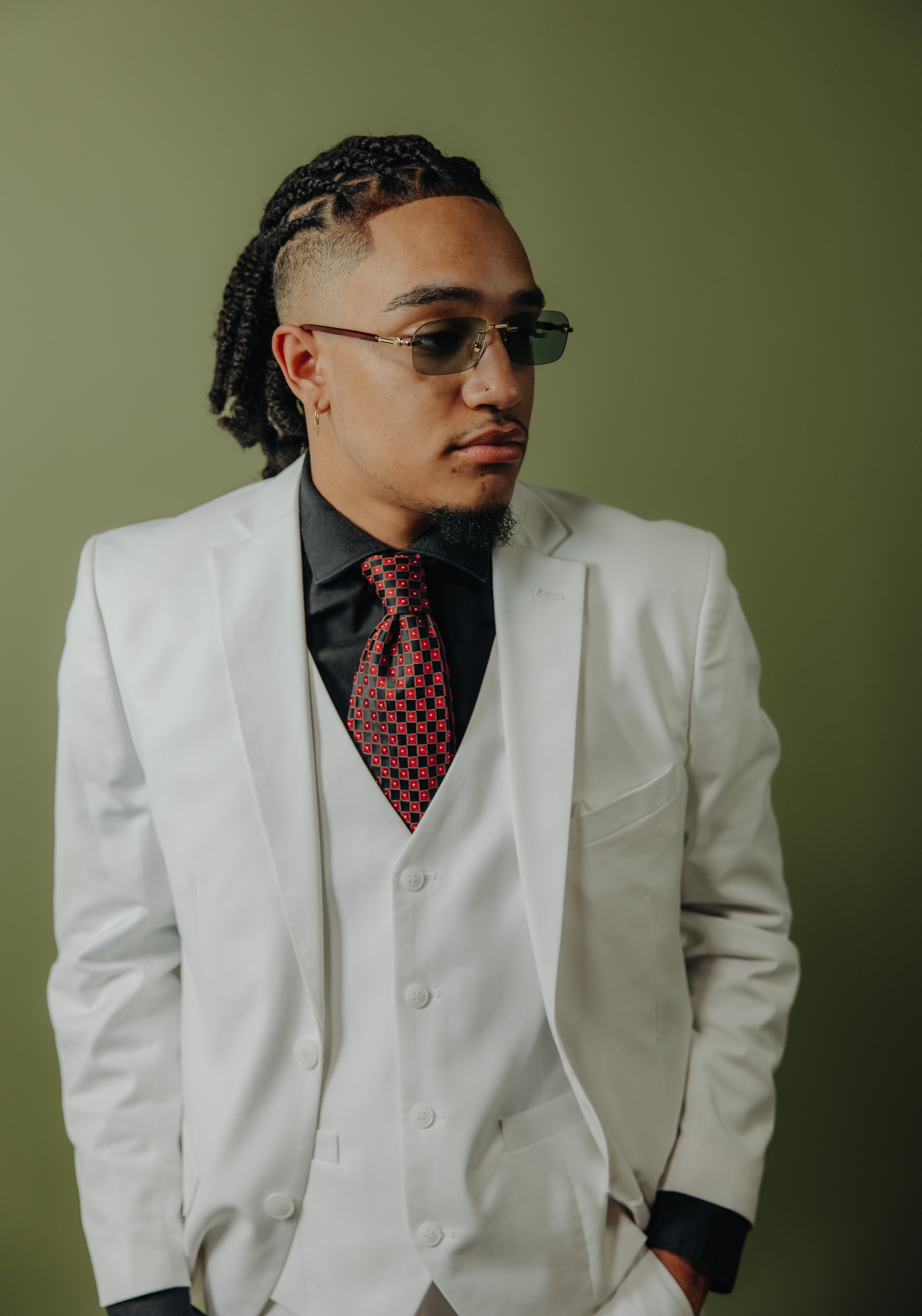 Man wearing a white suit with a black shirt and patterned red tie, sunglasses, and braided hairstyle, standing against a green background.
