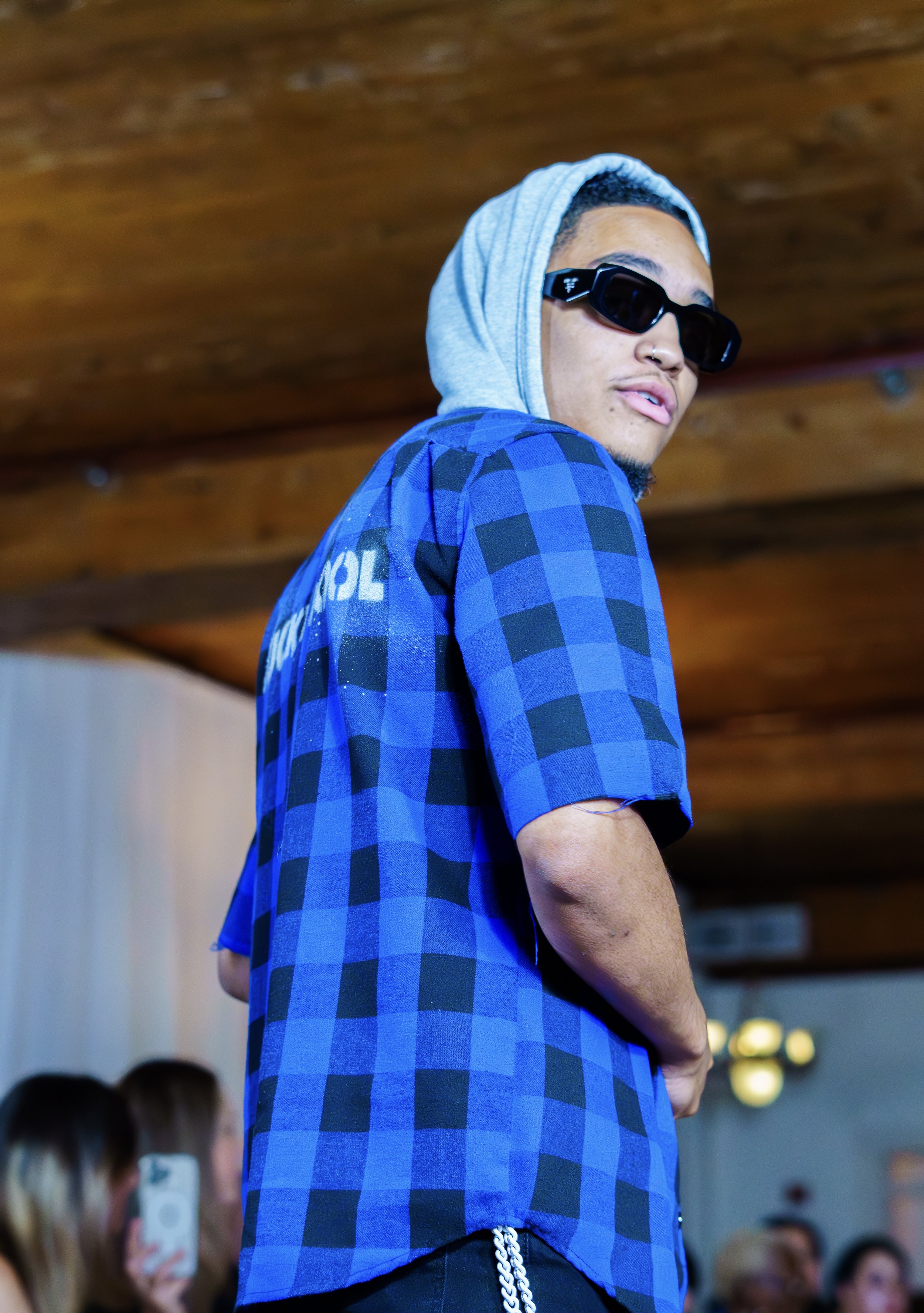 Person wearing sunglasses, a blue and black checkered shirt, and a gray hood, standing indoors.