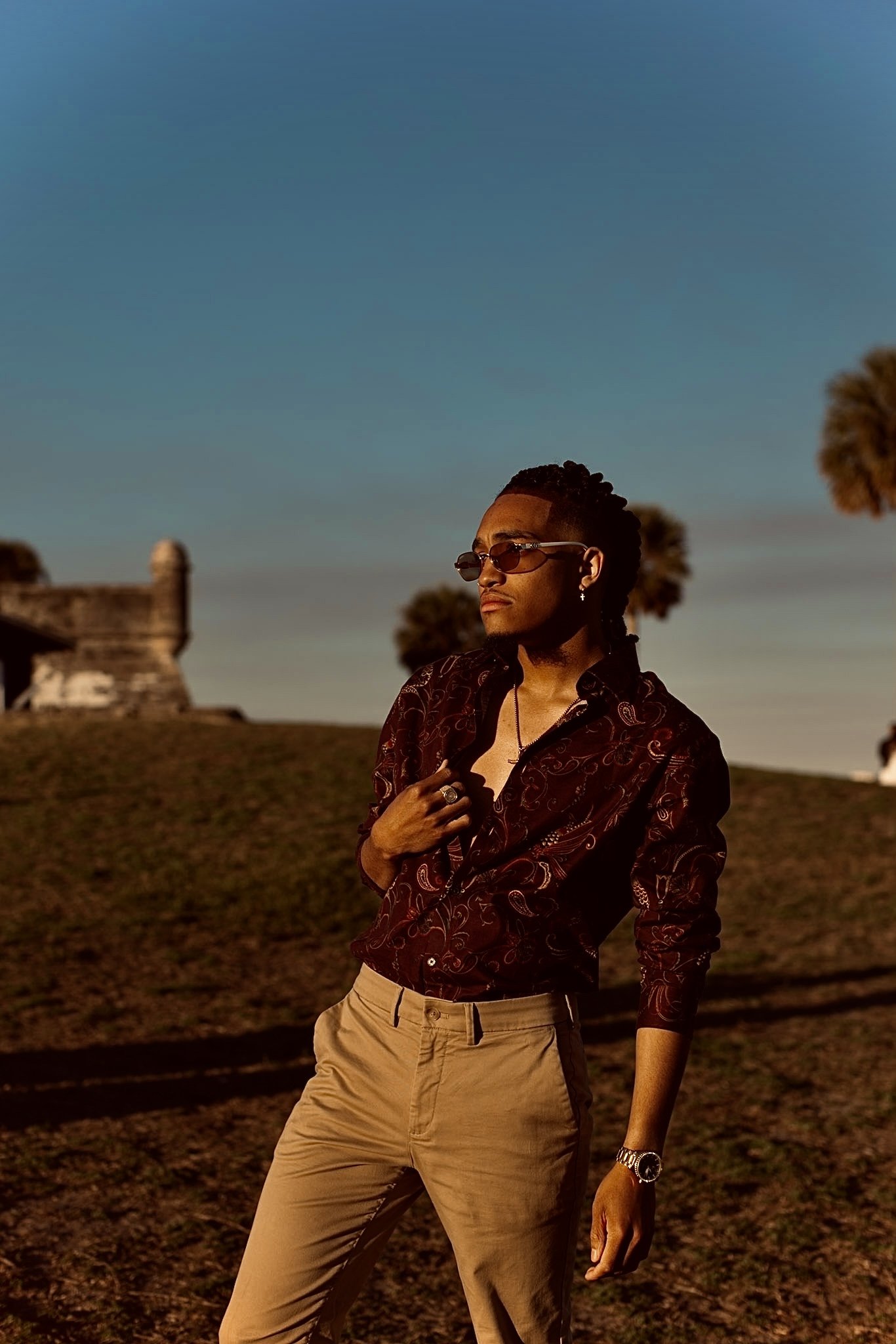 A person wearing sunglasses, a patterned shirt, and beige pants stands outdoors on a grassy hill with a blue sky and trees in the background.