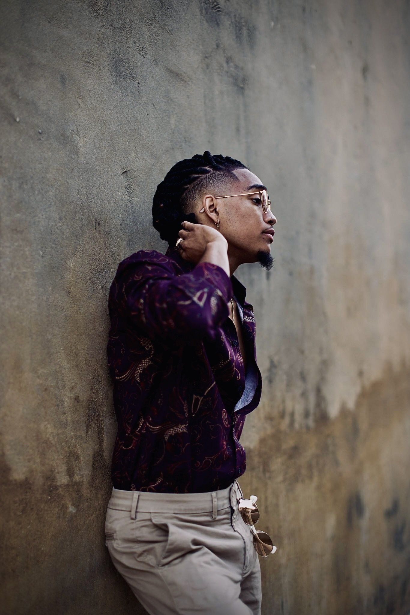 Man with braided hair leaning against a wall, wearing a patterned shirt, glasses, and beige pants with sunglasses hanging from the pocket.