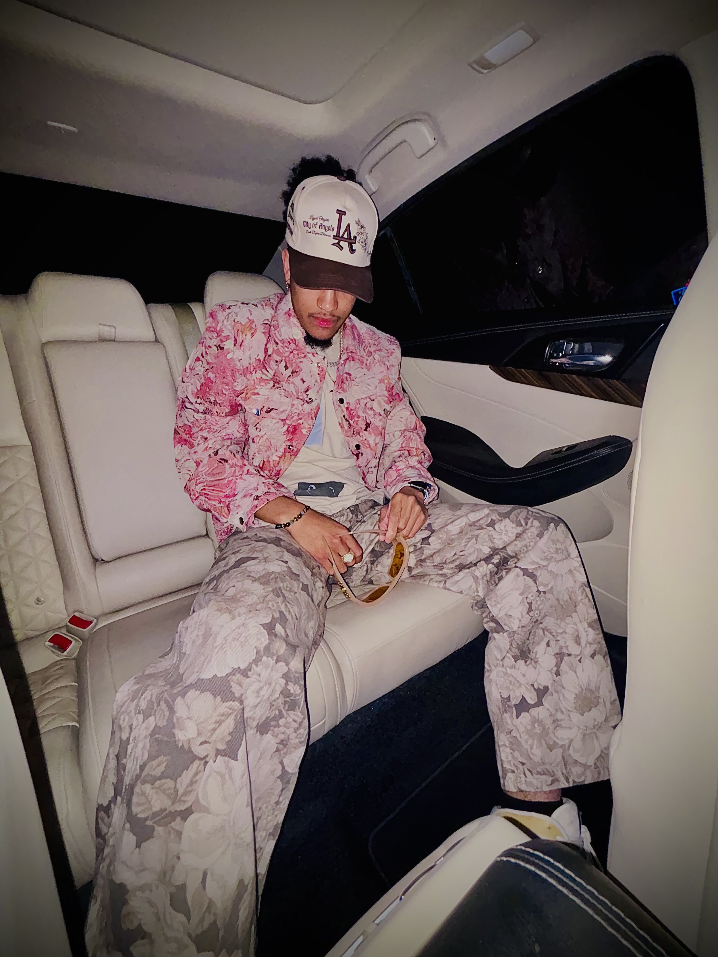 Person wearing a floral-patterned jacket and pants sitting in a car. They have a cap with the letters "LA" on it and are holding a belt.