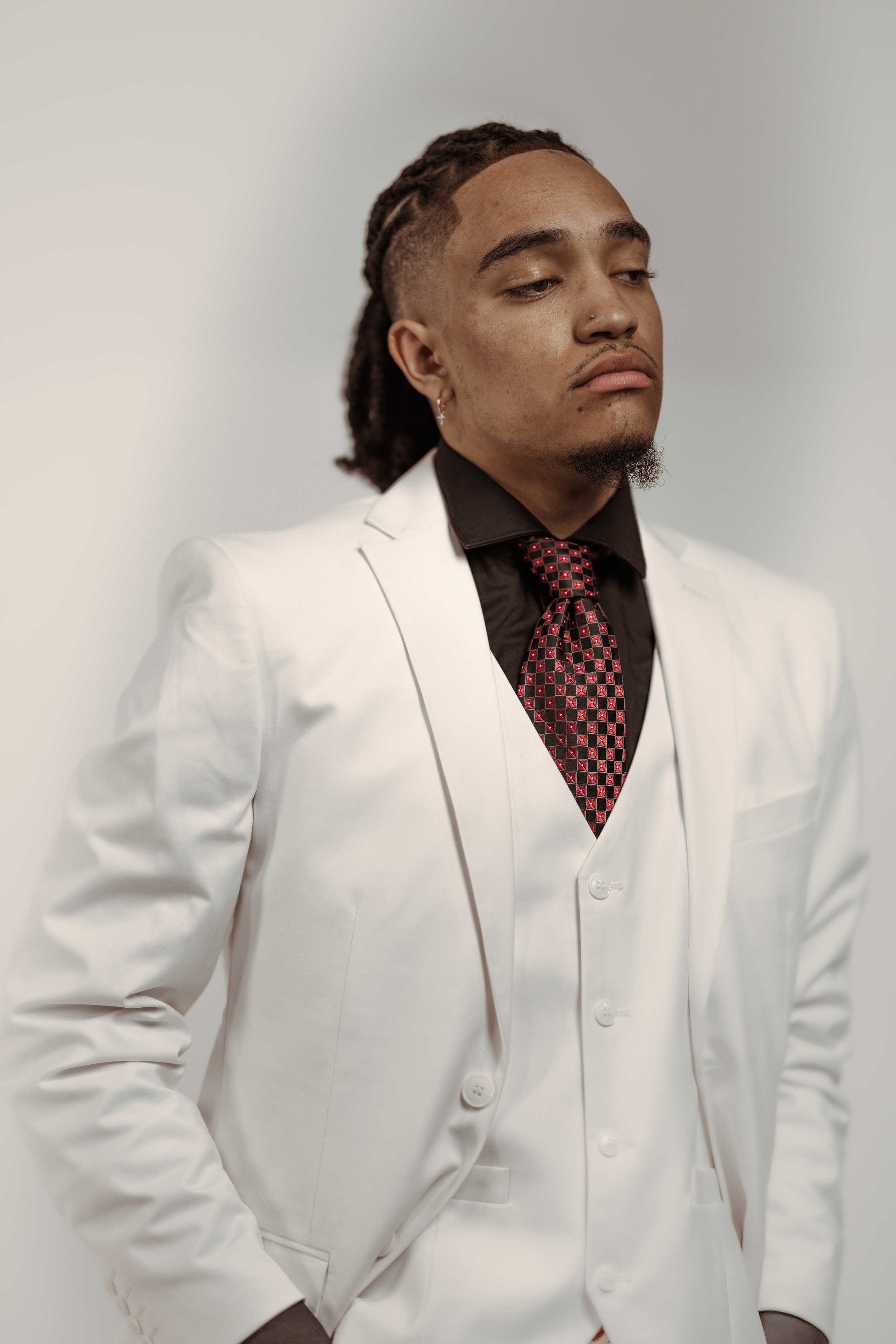 Person wearing a white suit and black shirt with a red and black tie, standing against a plain background.