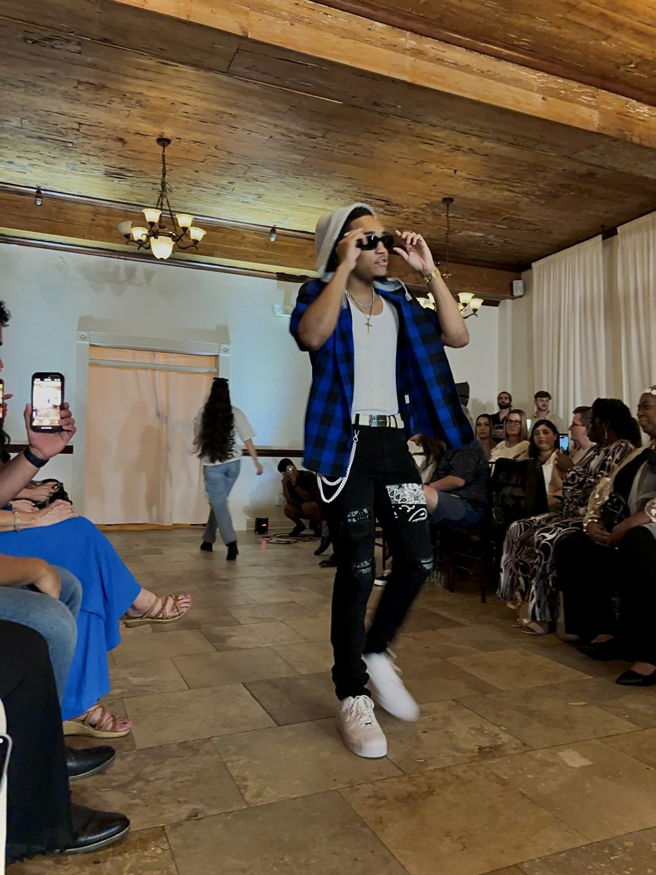 Model walking on a runway during a fashion show, wearing a blue flannel shirt, white tank top, ripped jeans, and a hat, with audience seated on either side.