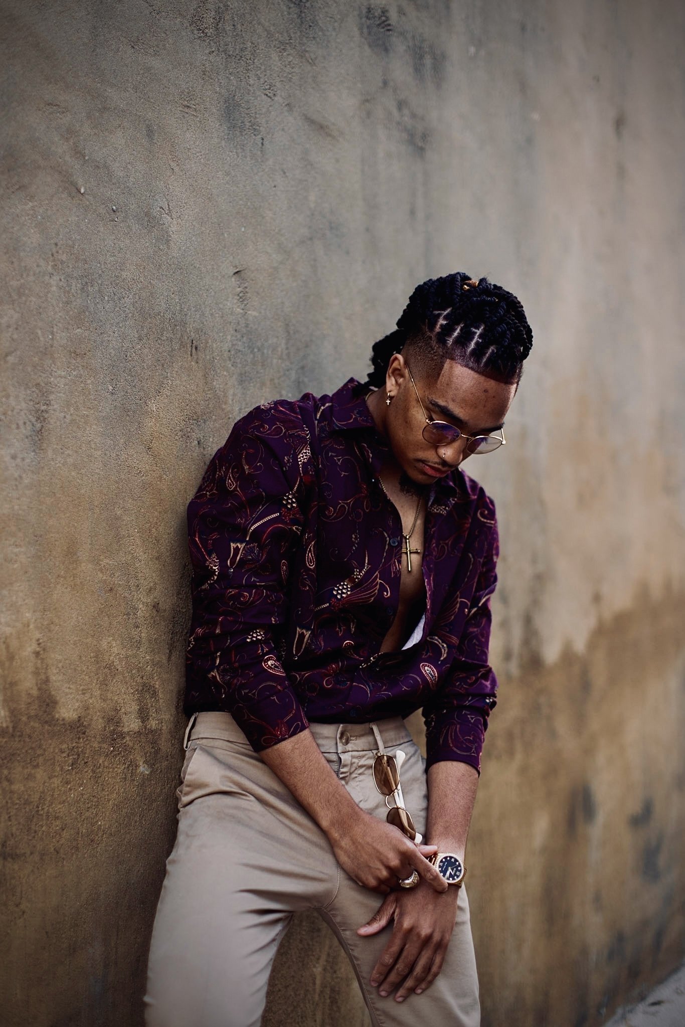 A man with braided hair wearing a patterned shirt, beige pants, and sunglasses, leaning against a textured wall.