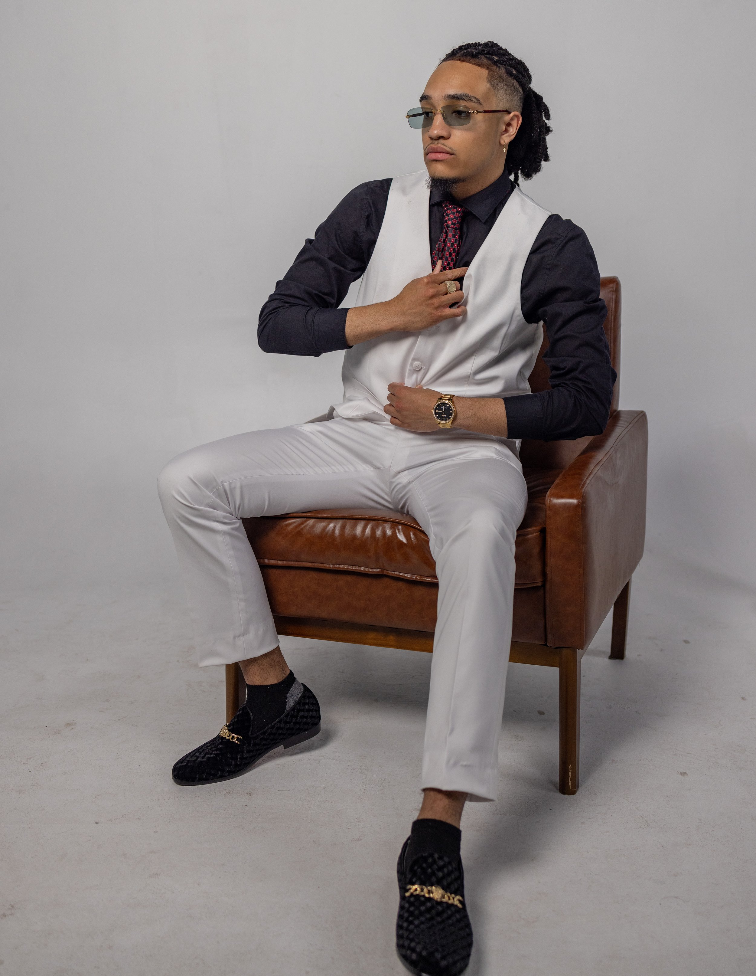 A stylishly dressed man sitting on a brown leather chair, wearing sunglasses, a black shirt, white vest, white pants, and black loafers with gold accents.