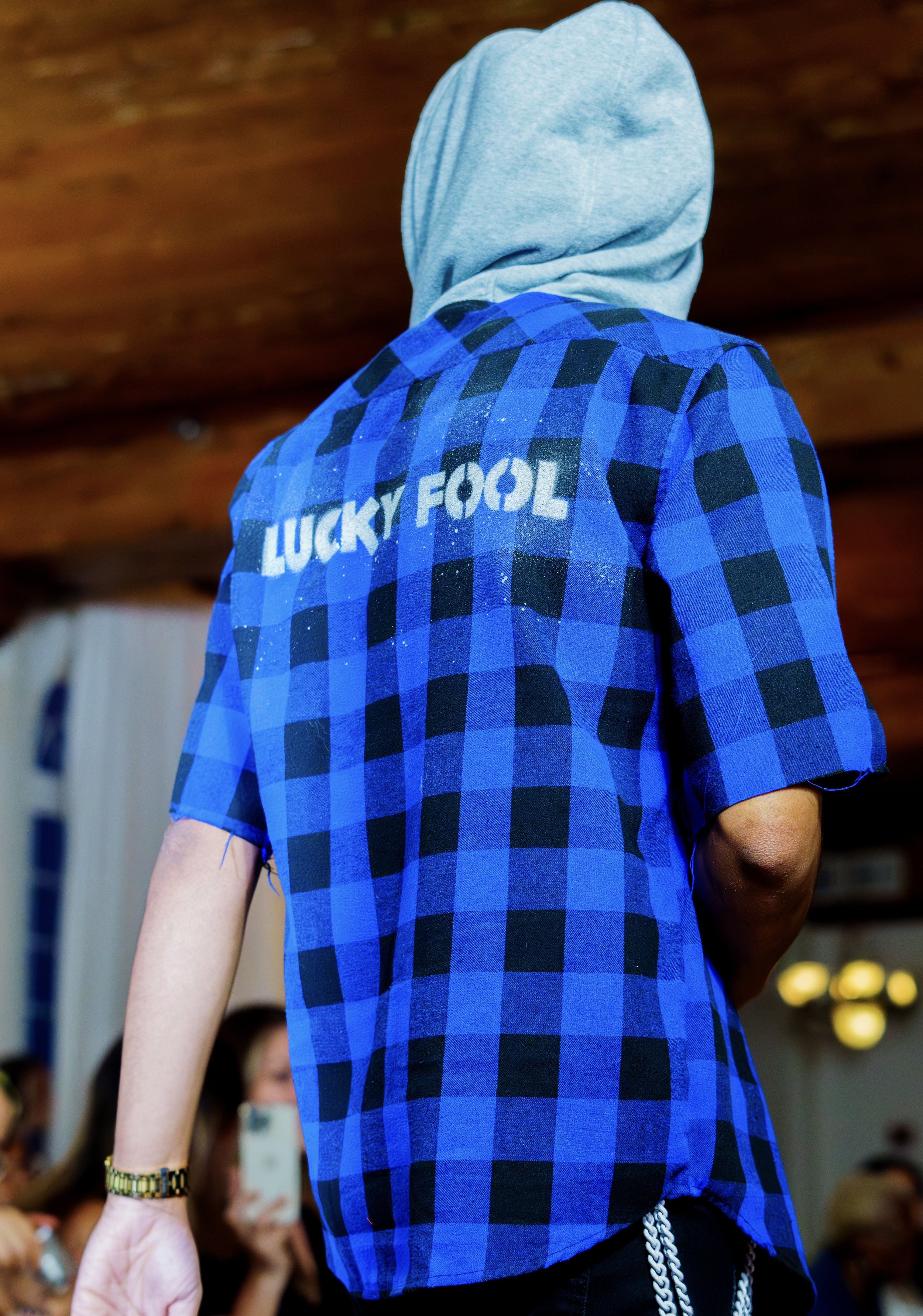 Back view of a person wearing a blue and black checkered shirt with 'LUCKY FOOL' text on the back, and a gray hood, indoors.