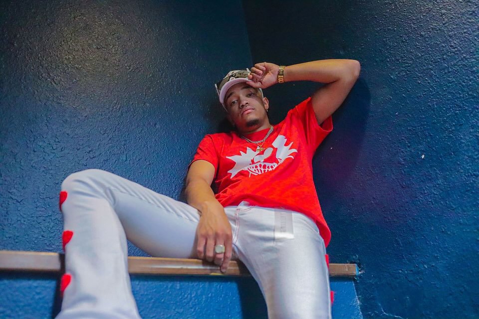 Person wearing a red t-shirt and white pants sitting against a blue wall, with one arm resting on their head.