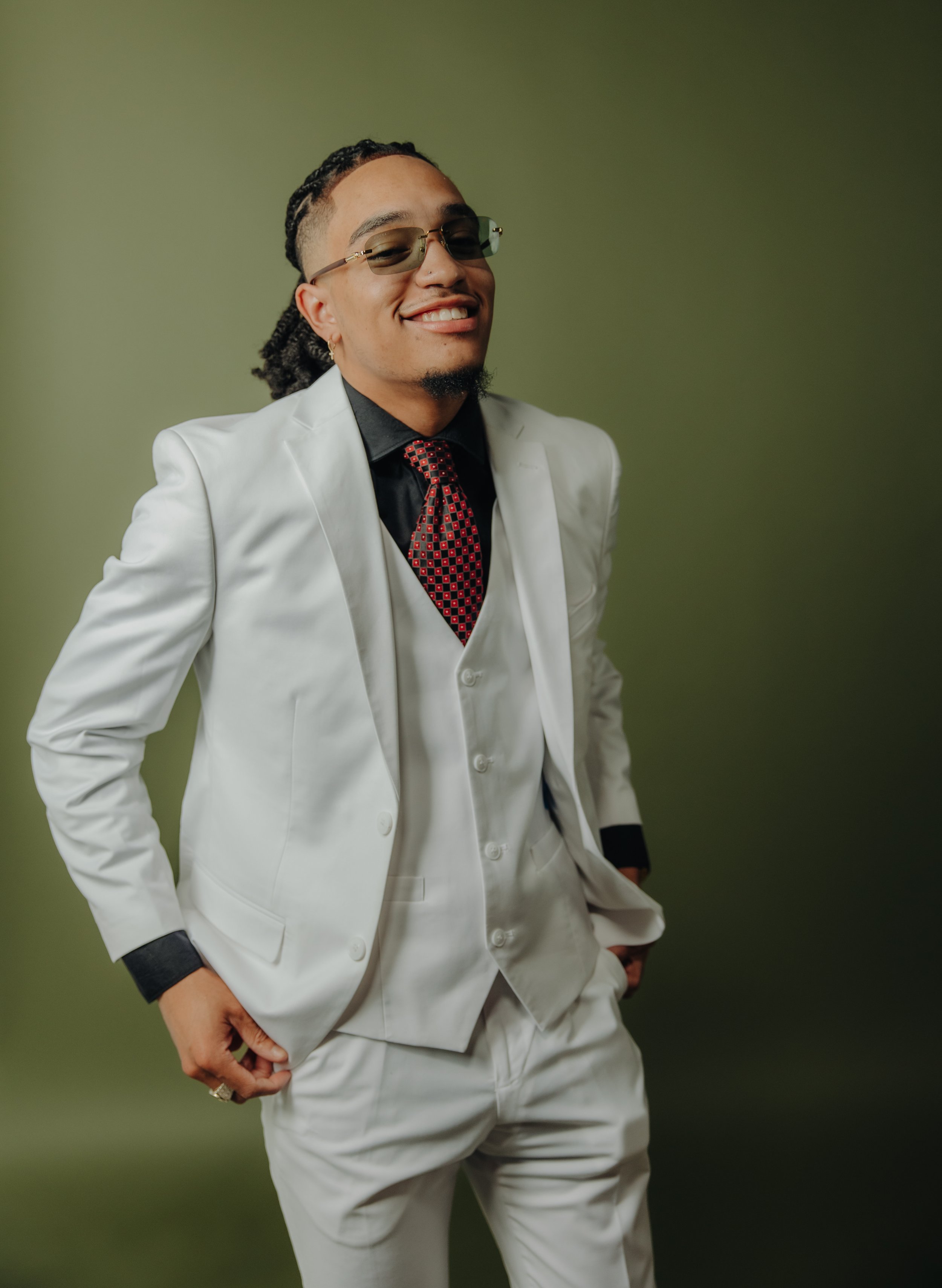 Person in a white suit and red patterned tie, wearing sunglasses, smiling against a green background.