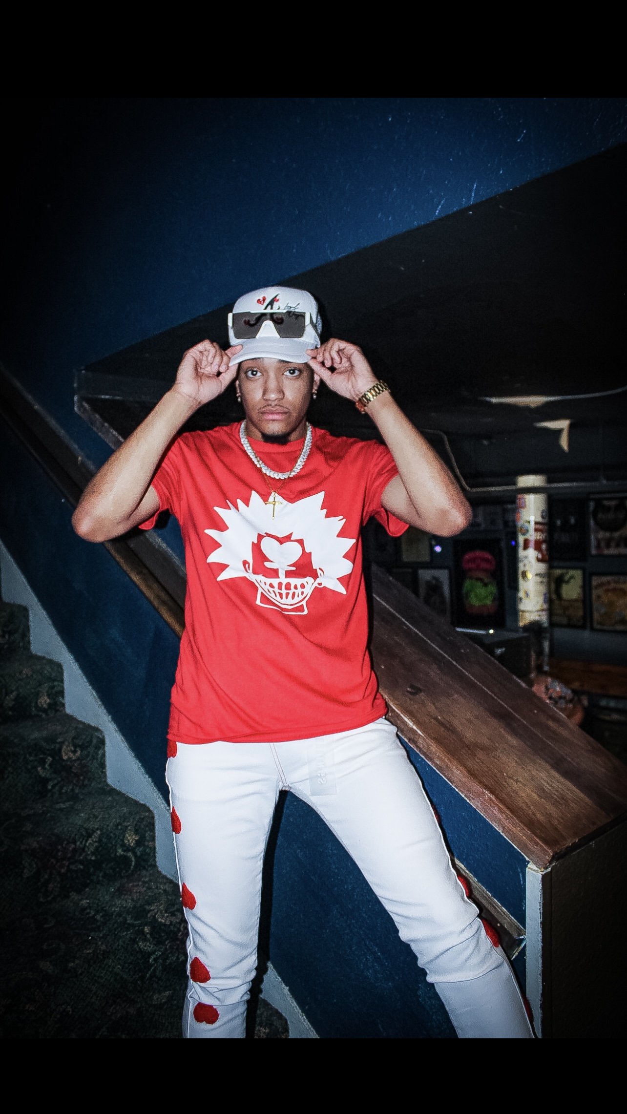 Person in a red shirt with a graphic design, white pants, and a white cap indoors.