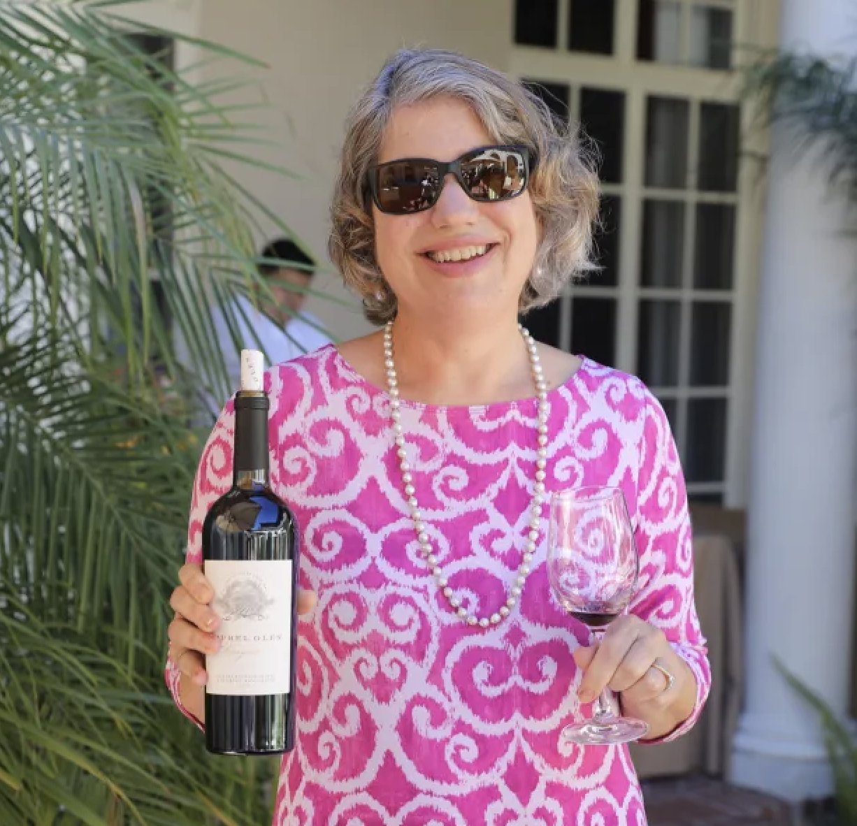 A smiling woman with curly gray hair wearing sunglasses, a pink and white patterned dress, a pearl necklace, and holding a bottle of red wine and a wine glass with red wine. Luxury Restaurant and Hotel in Bucks County Pennsylvania