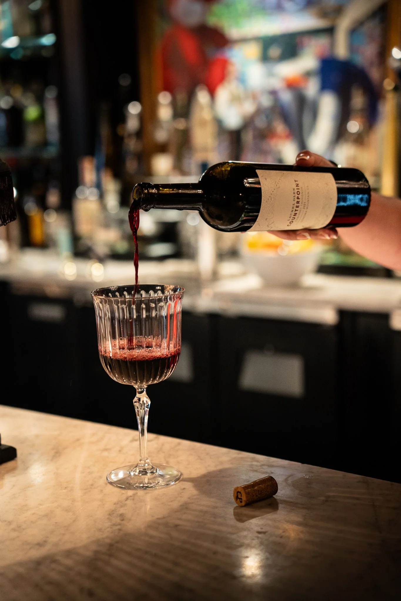 A person is pouring red wine from a bottle into a crystal wine glass on a marble countertop. A cork is on the counter nearby, and the background shows a blurred bar area with various bottles and colorful decor. Luxury Restaurant and Hotel in PA