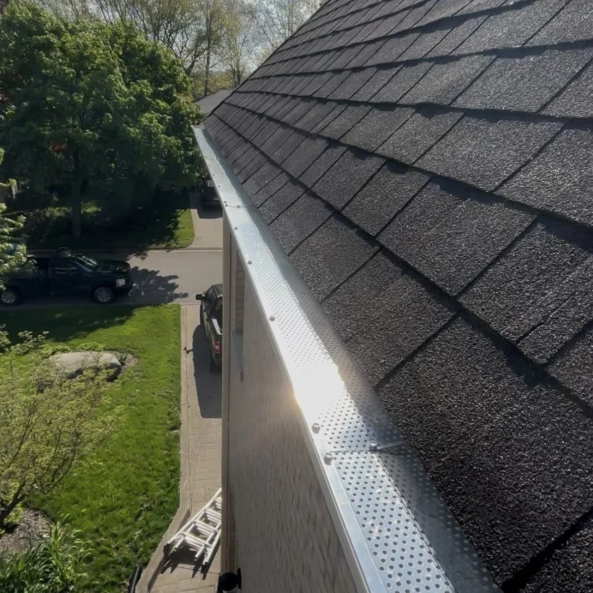 View from a ladder showing a house roof with black shingles and a metal gutter. Trees, parked cars, and a street are visible below.