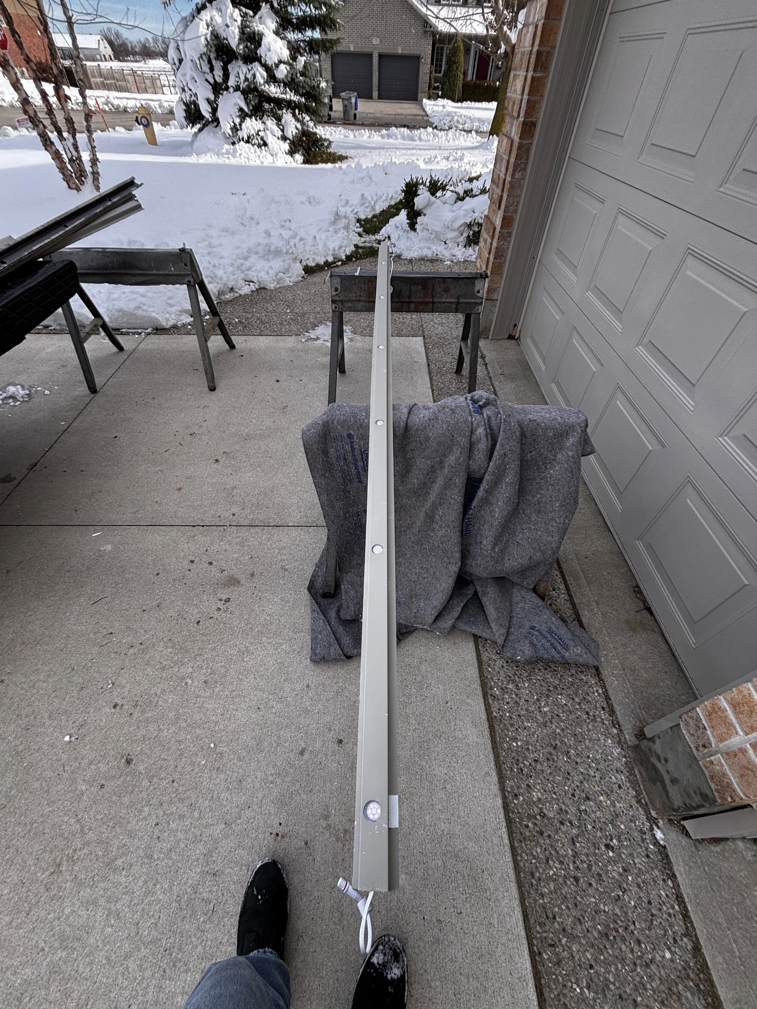 A person is standing on a concrete porch holding a long metal piece, with snow and trees in the background, and a partially disassembled object on sawhorses.