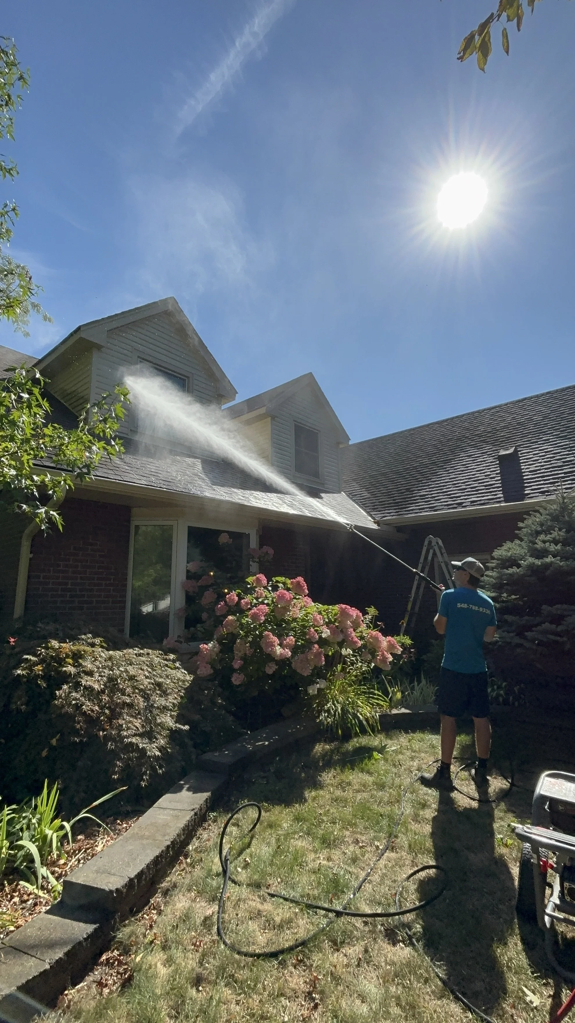 A person, wearing a blue shirt and gray cap, is pressure washing the roof of a house on a sunny day. The house has a brick exterior, pink flowering bushes, and is surrounded by greenery.