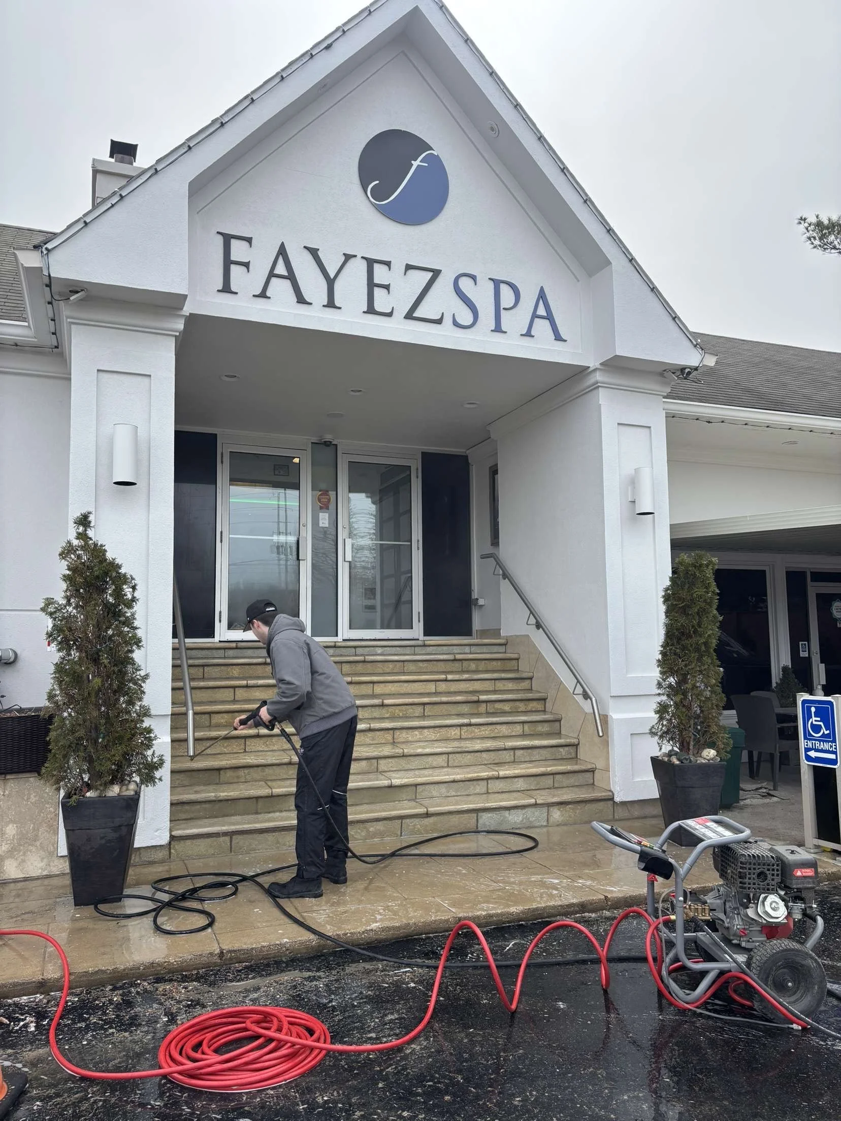 Person power washing the steps outside Fayez Spa building with a pressure washer machine on a wet surface, near potted plants and a handicap accessible sign.