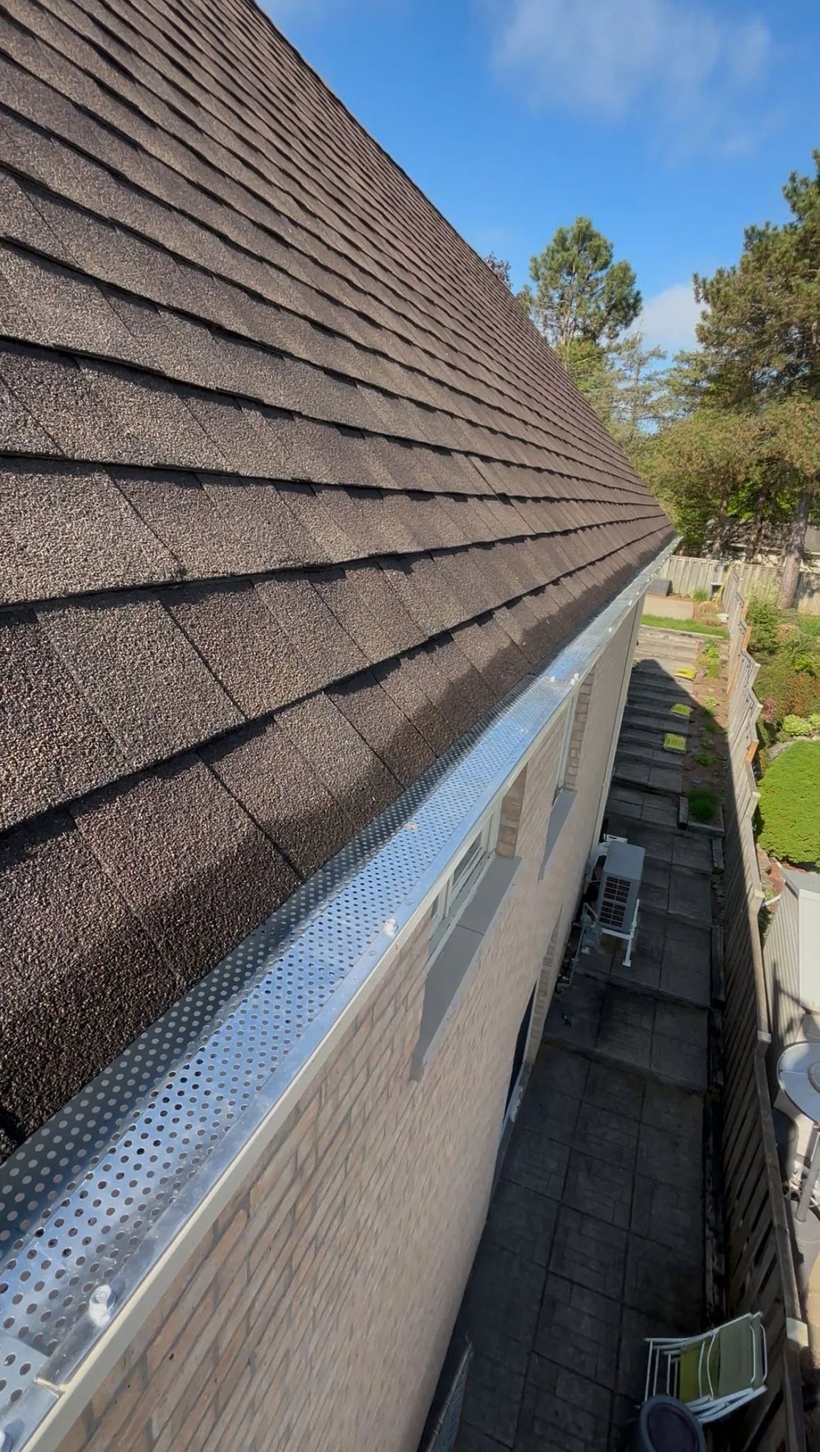 View of a house roof with brown shingles and a metal drip edge, along with part of a backyard, fence, trees, and a blue sky.