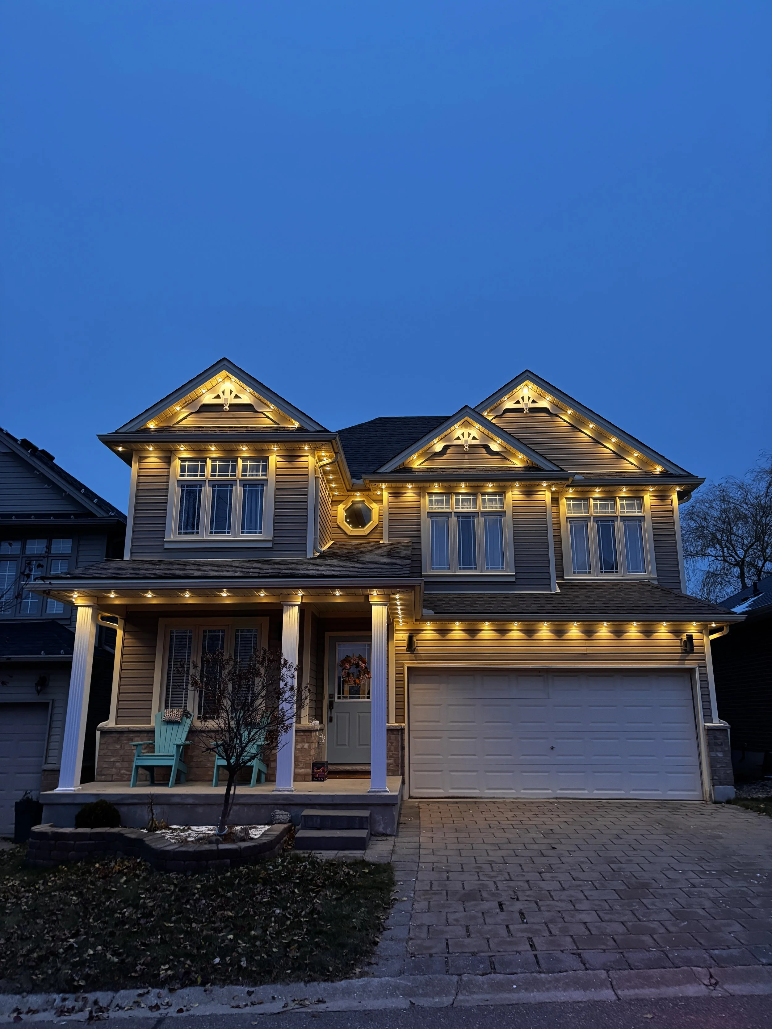 A two-story house decorated with yellow Christmas lights along the roofline and porch, with a small front yard and driveway at dusk.