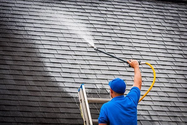 A man wearing a blue shirt and matching cap power washing a house roof with a yellow hose and spray wand.