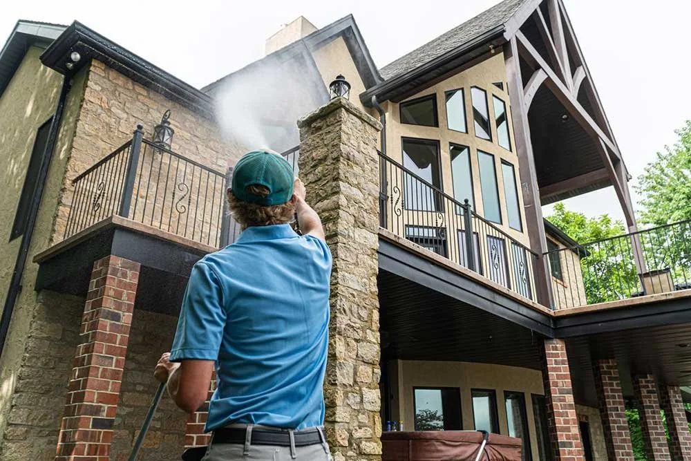 A person wearing a blue shirt and green cap is pressure washing the exterior of a multi-story house with stone, brick, and wood siding, with smoke or spray coming from the high-pressure washer.