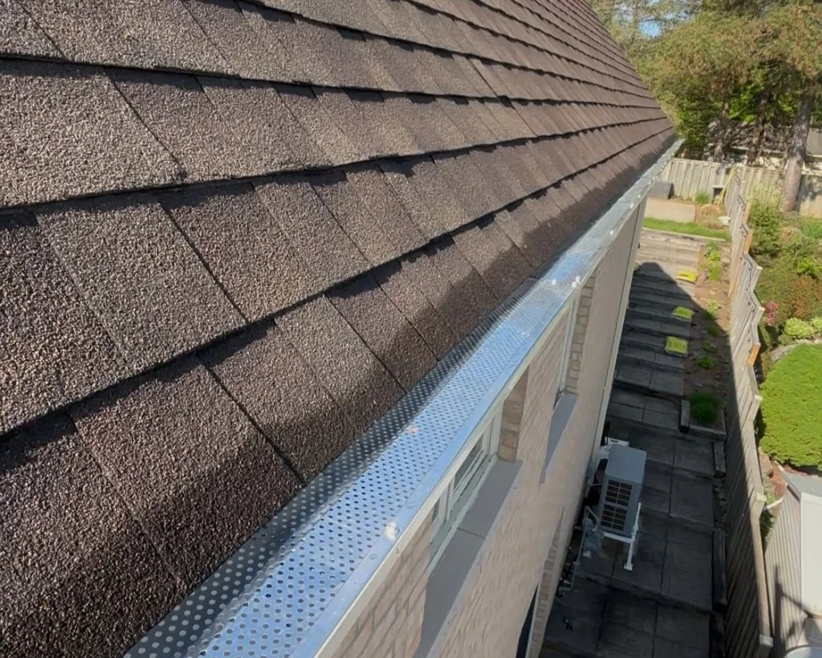 Close-up view of a house roof with brown asphalt shingles and a metal gutter along the edge, overlooking a backyard with a wooden fence, trees, and garden beds.