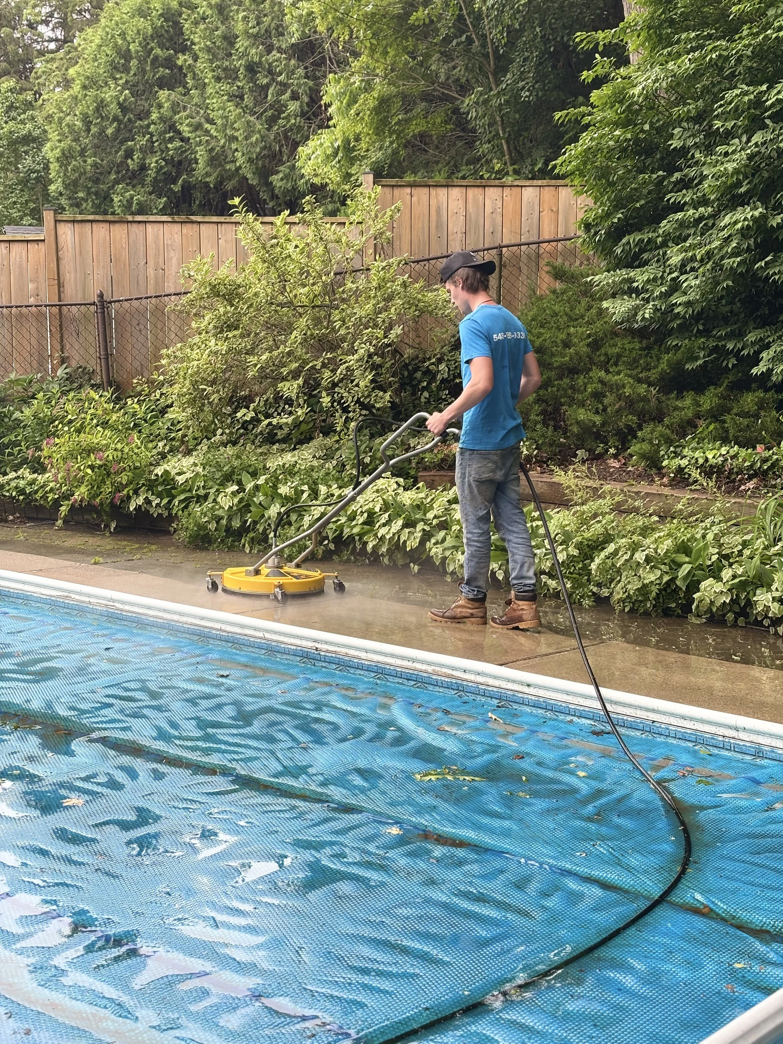 A person cleaning a swimming pool using a yellow pool cleaning machine, with green bushes and a wooden fence in the background.