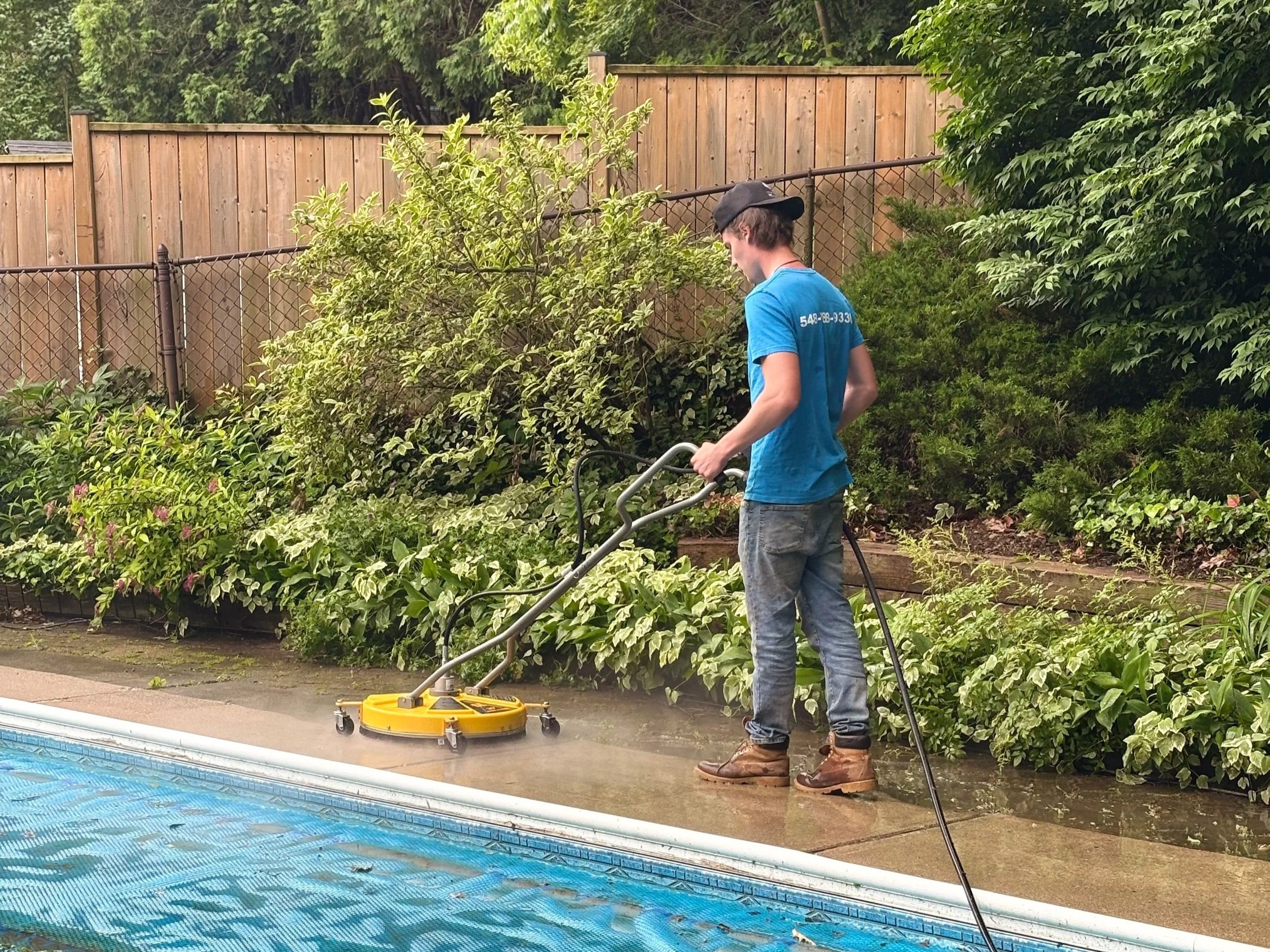 A man in a blue t-shirt and jeans is using a yellow pool vacuum to clean the pool deck near the pool, which is covered with a blue tarp, in a backyard with green bushes and a wooden fence.