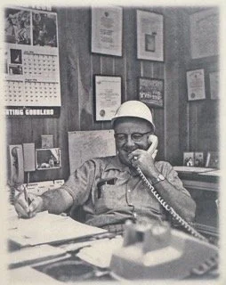 A man sitting at a desk on the phone in an office with framed pictures and certificates on the wall behind him.
