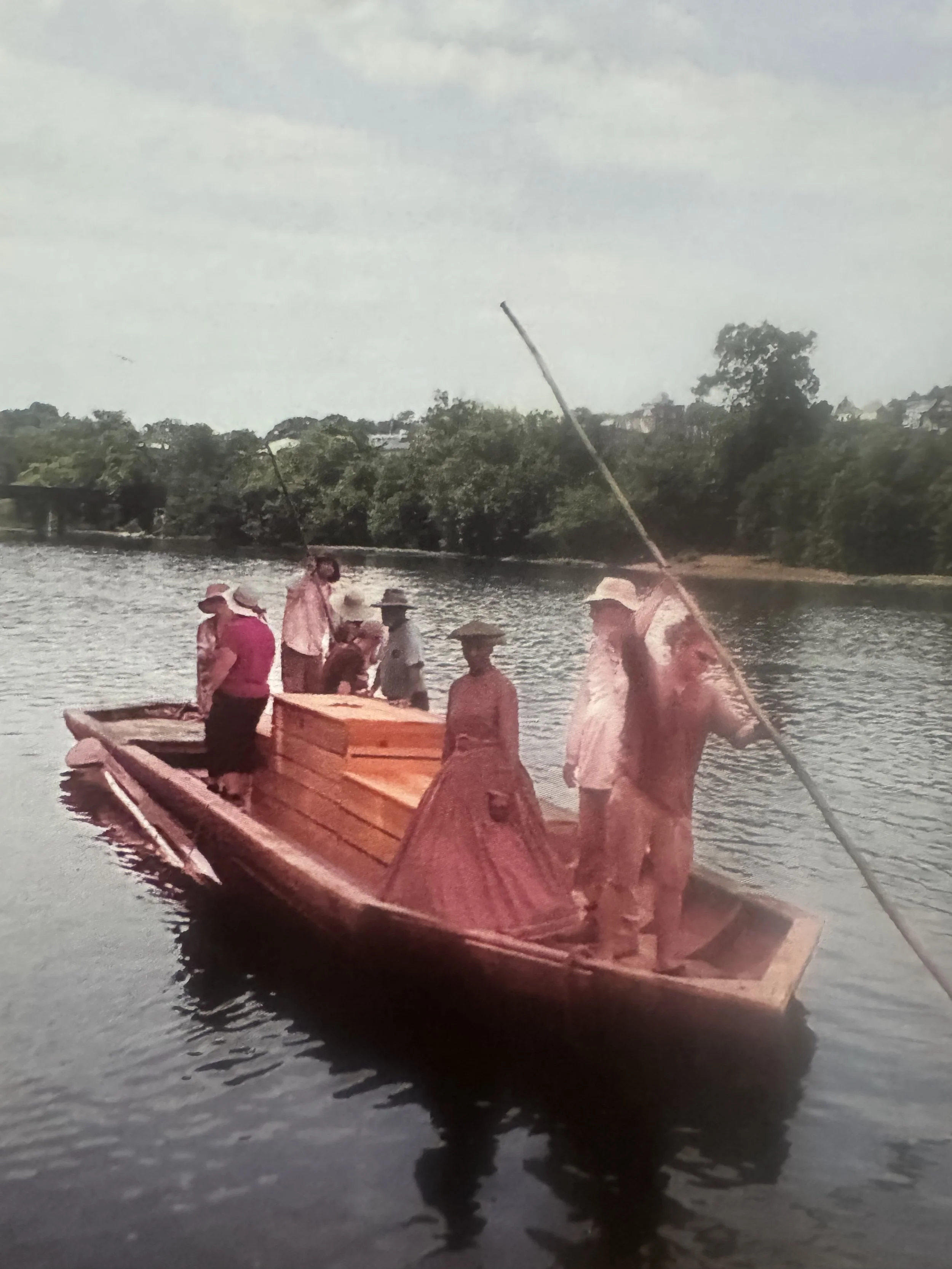 Several people on a wooden boat in a river, some holding long poles, with trees in the background.