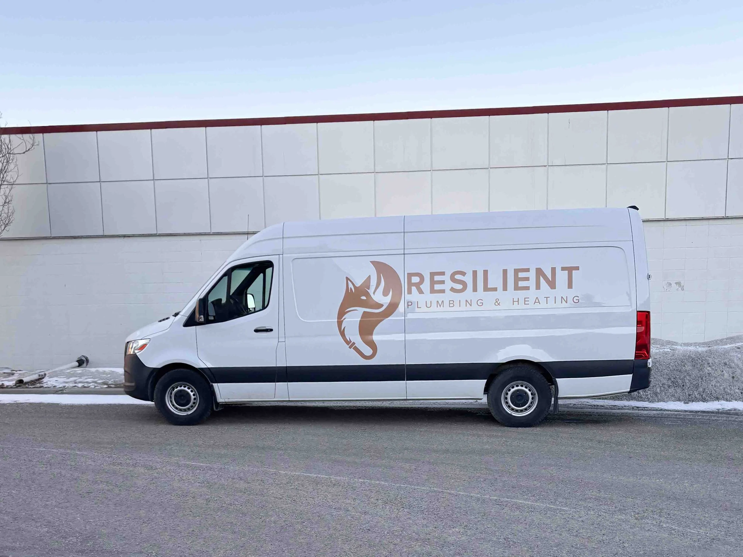 White service van with logo and text for Resilient Plumbing & Heating parked on road in winter, with snow and a plain white building in background.