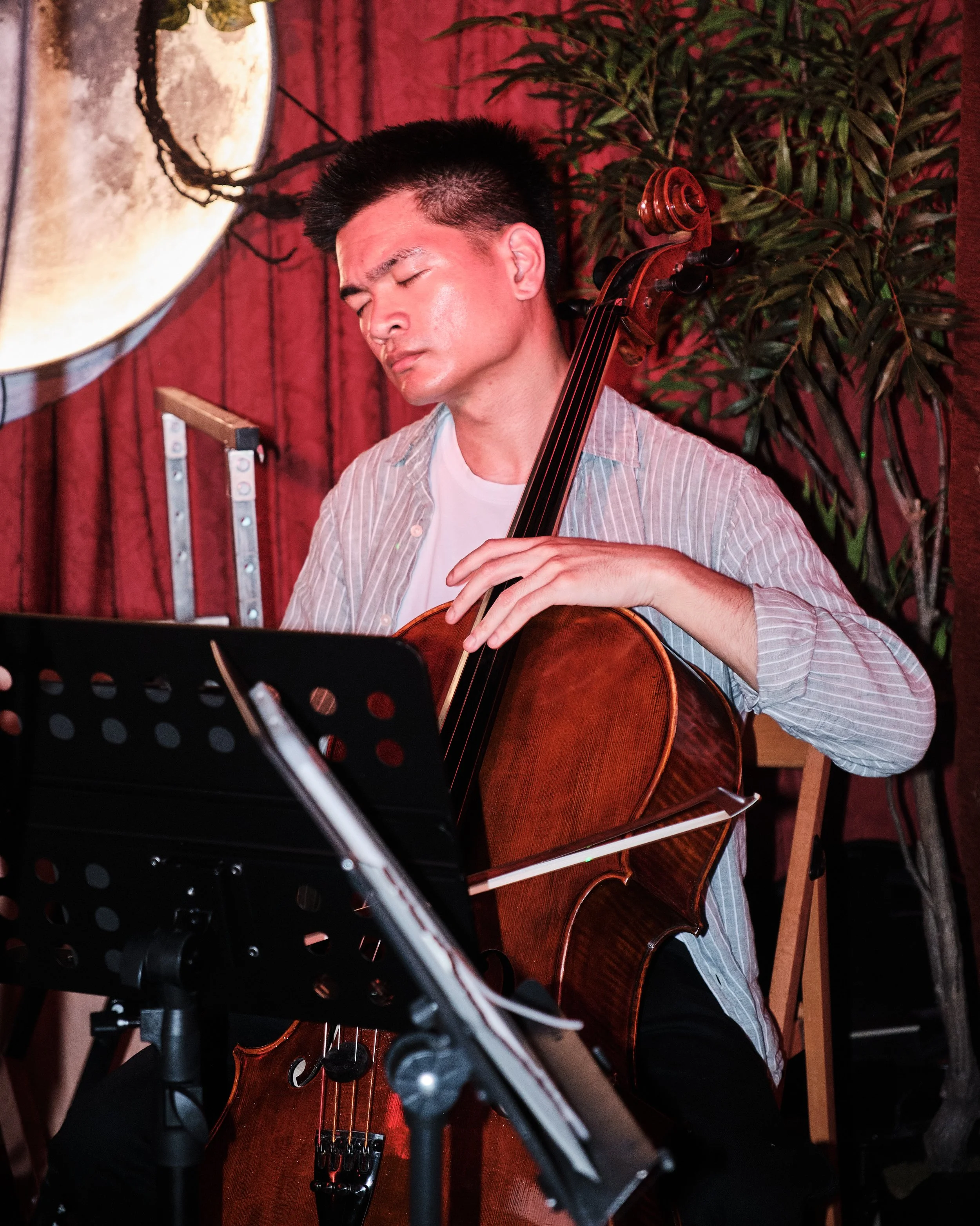 A person playing the cello indoors with music sheets nearby, surrounded by musical equipment.