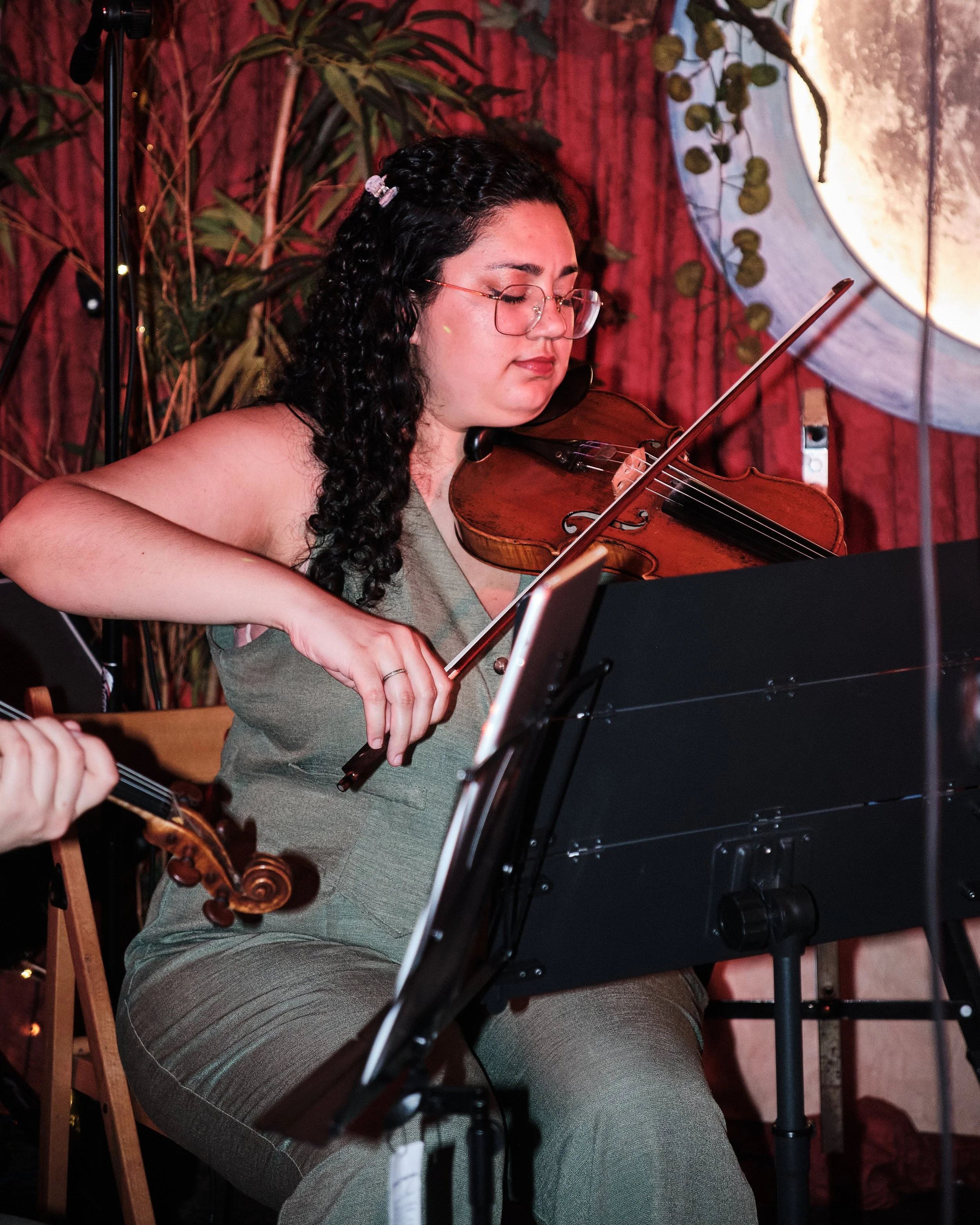 A woman playing a violin at a gathering with a lamp and wooden wall in the background, with others observing.